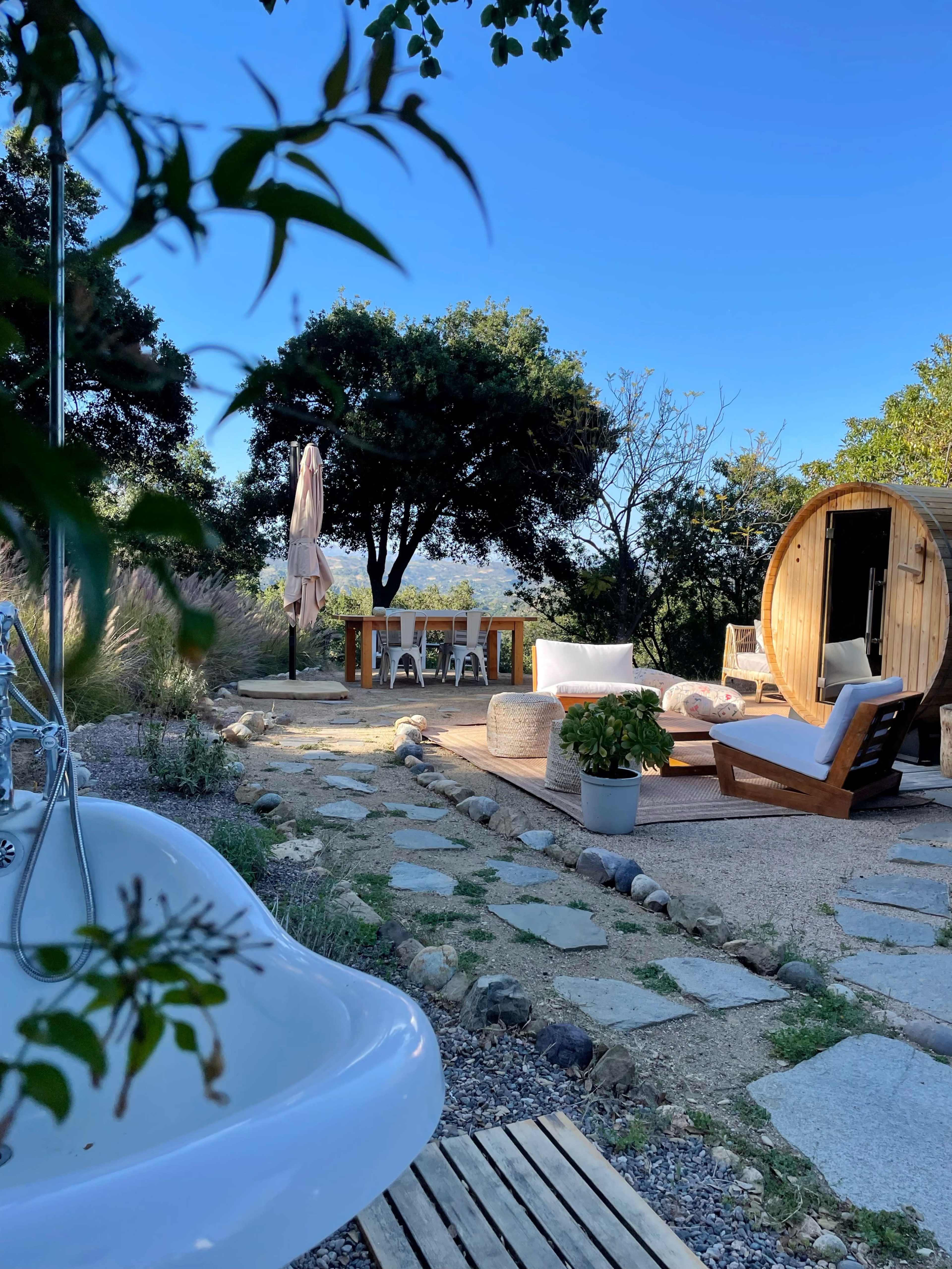 A wooden seating area and a dining table are set up outdoors near a bathtub and a barrel-shaped structure, all surrounded by trees and a clear blue sky.