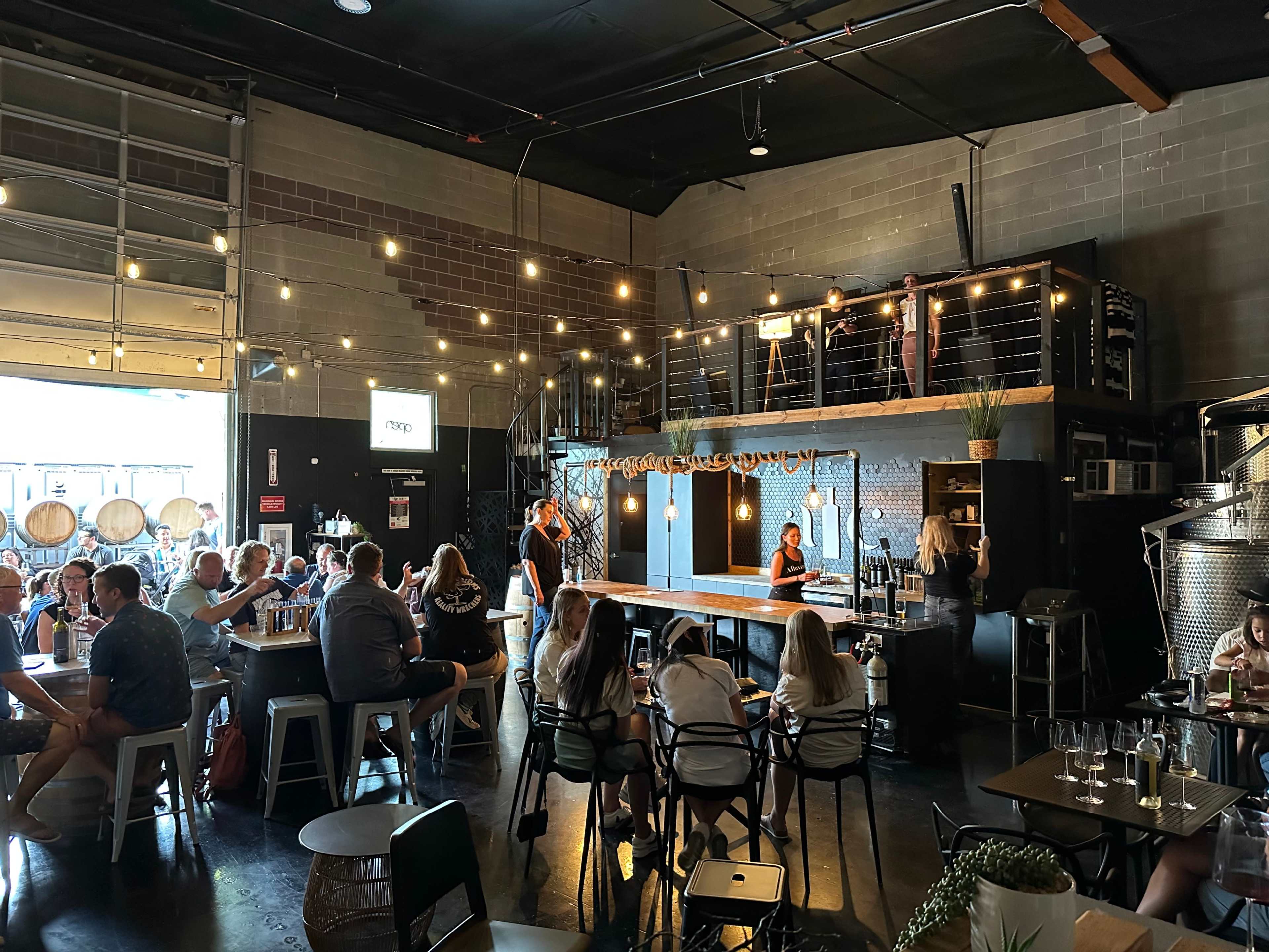 A crowded brewery features a bar area with patrons seated at tables, string lights hanging from the ceiling, and a two-level loft with additional seating.
