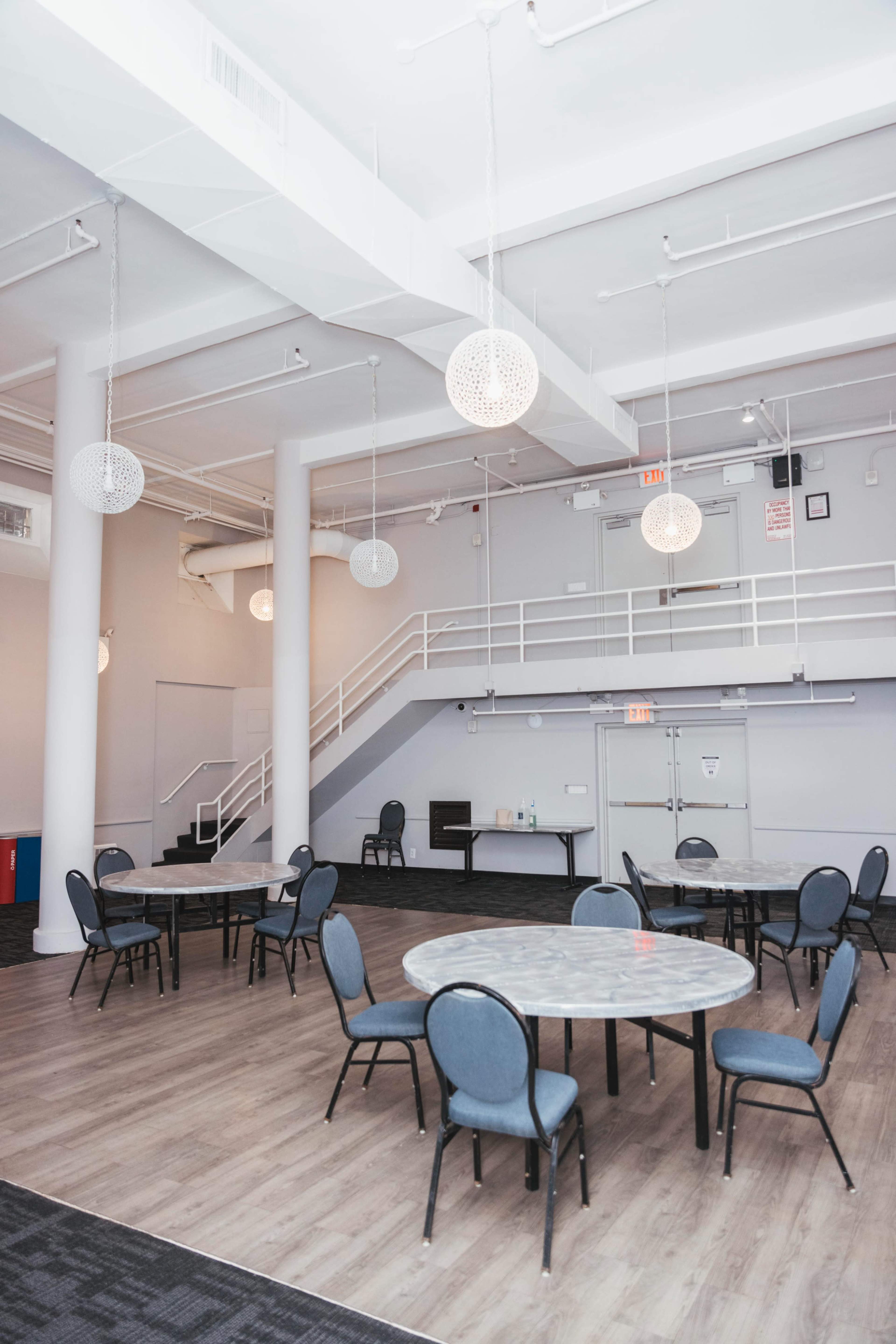 The image shows an empty event space with several round tables and chairs, modern light fixtures hanging from the ceiling, and a staircase in the background.