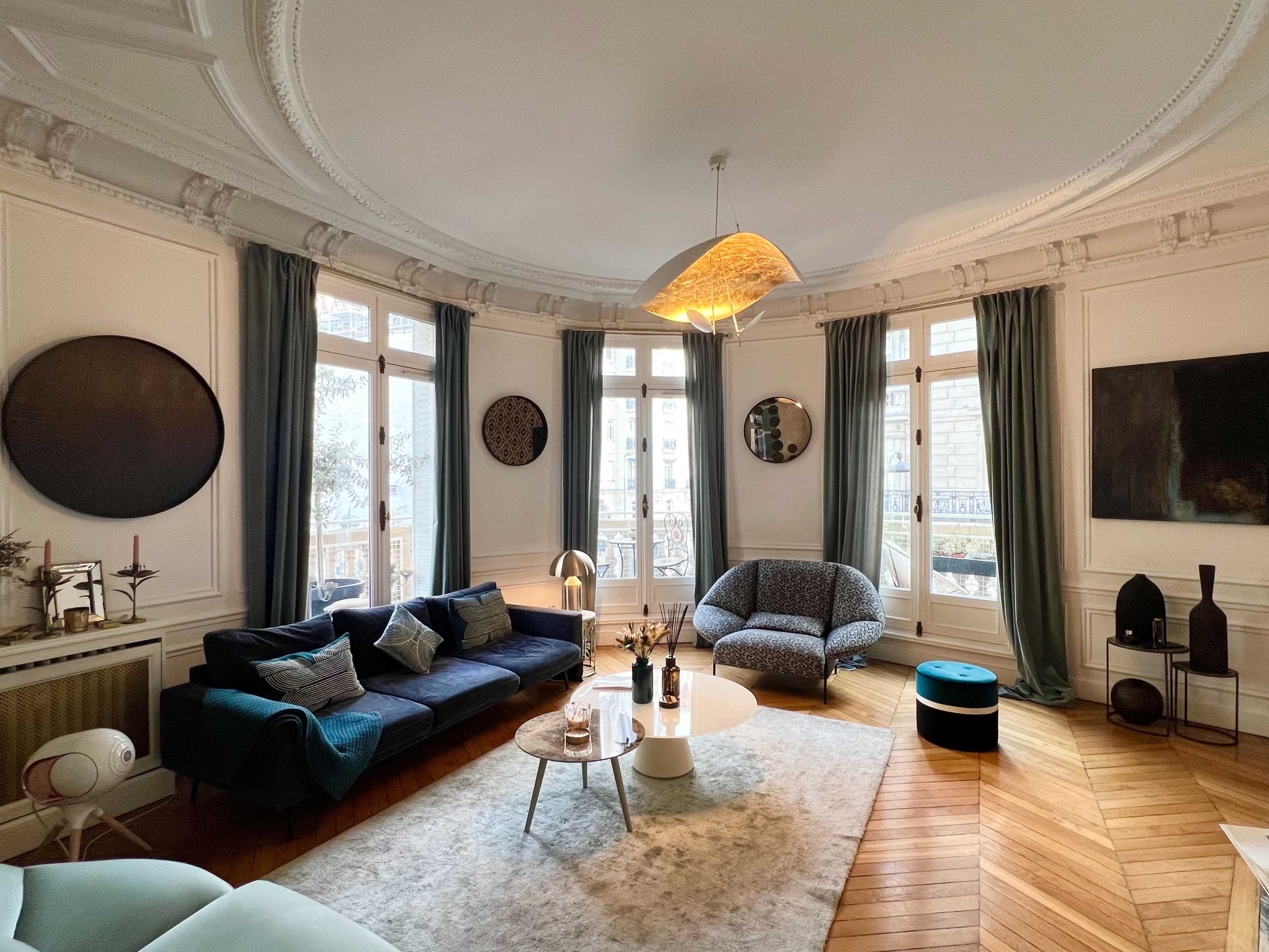 A stylish living room with a blue sofa, a round coffee table, a patterned armchair, and large windows allowing natural light to enter.