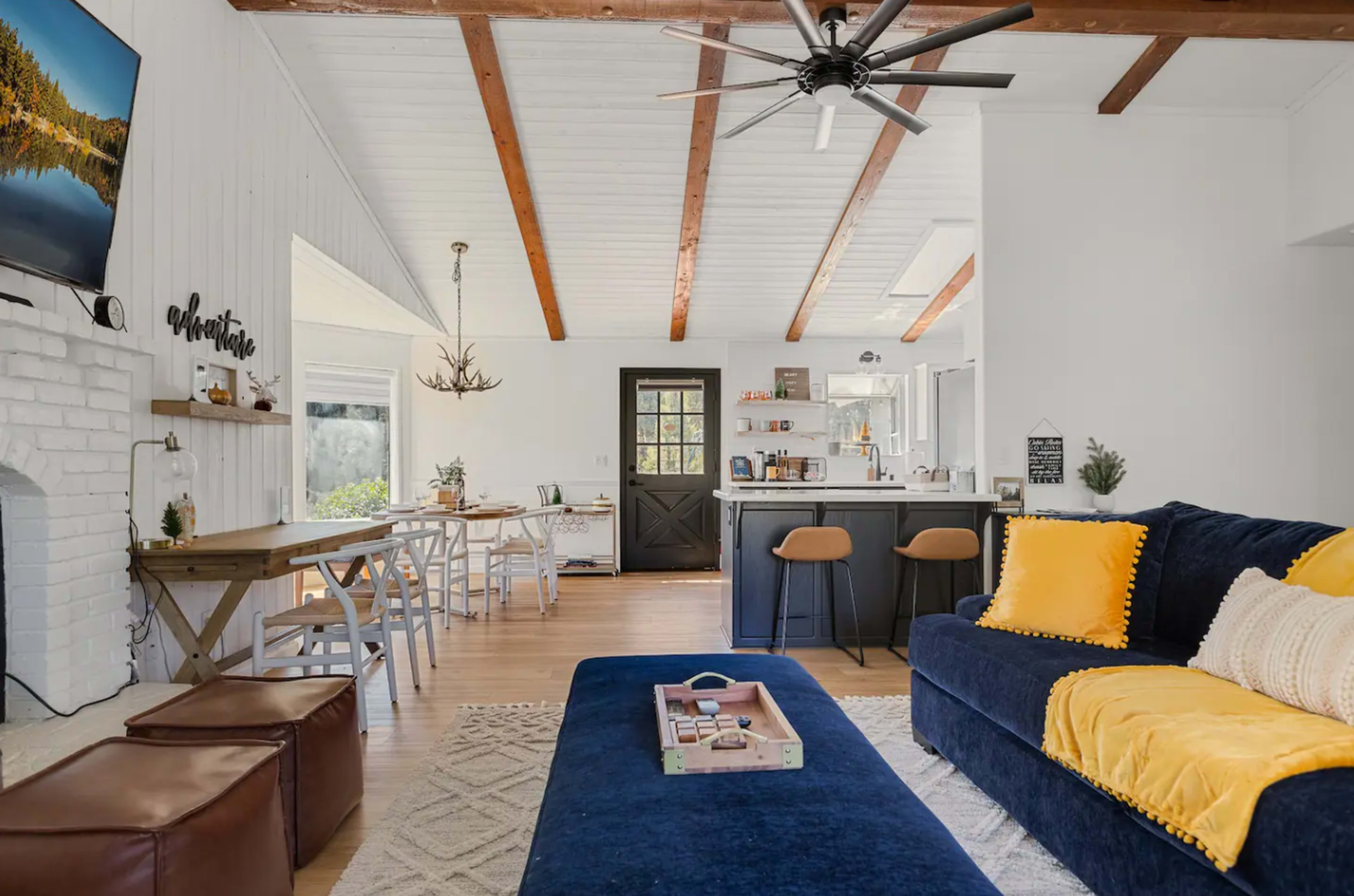 The image shows a modern living room with a blue sofa, a wooden dining table, and a kitchen area featuring a bar with stools.