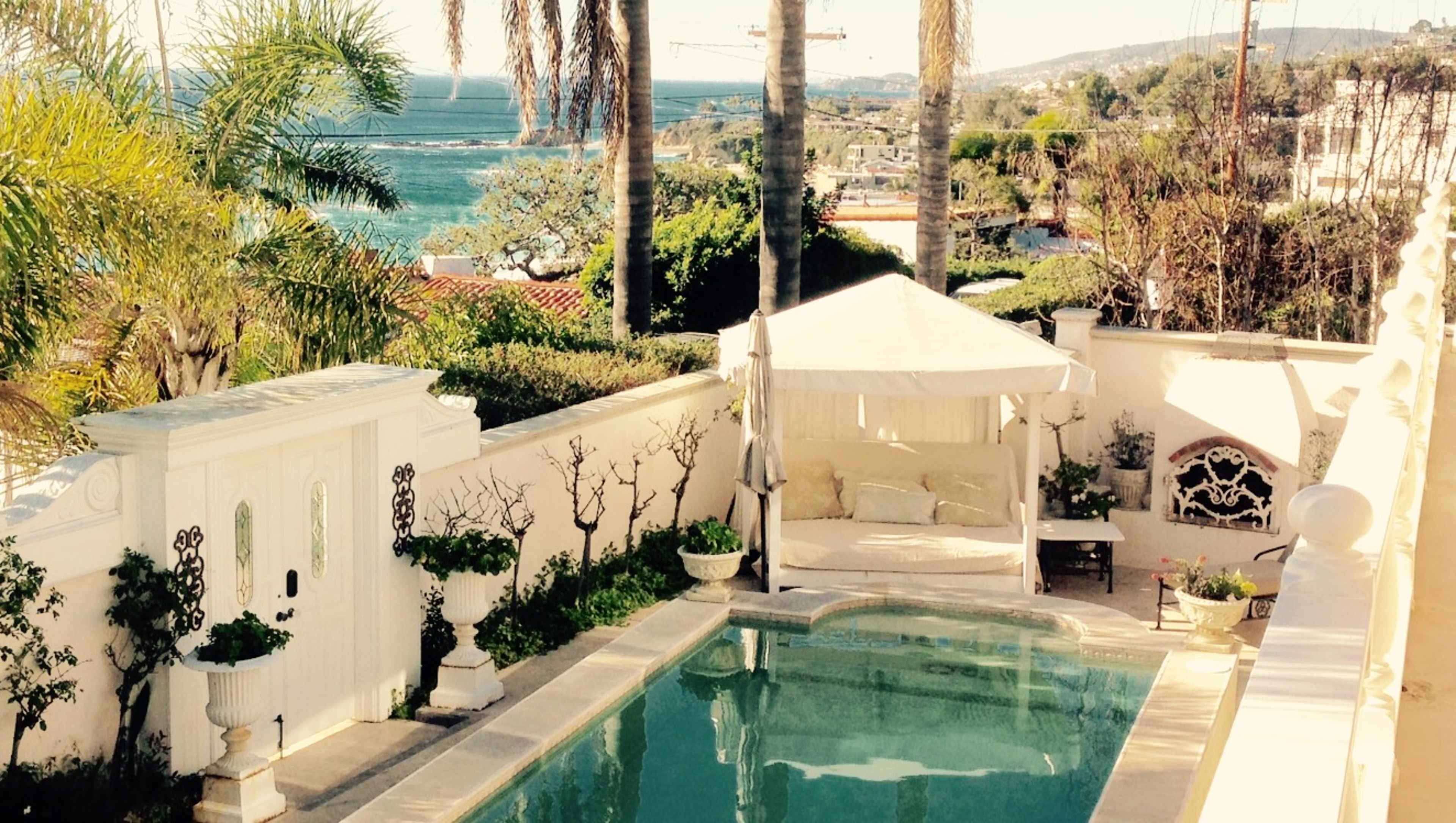 The image features a private pool surrounded by palm trees, a lounging area draped with fabric, and a view of the ocean in the background.