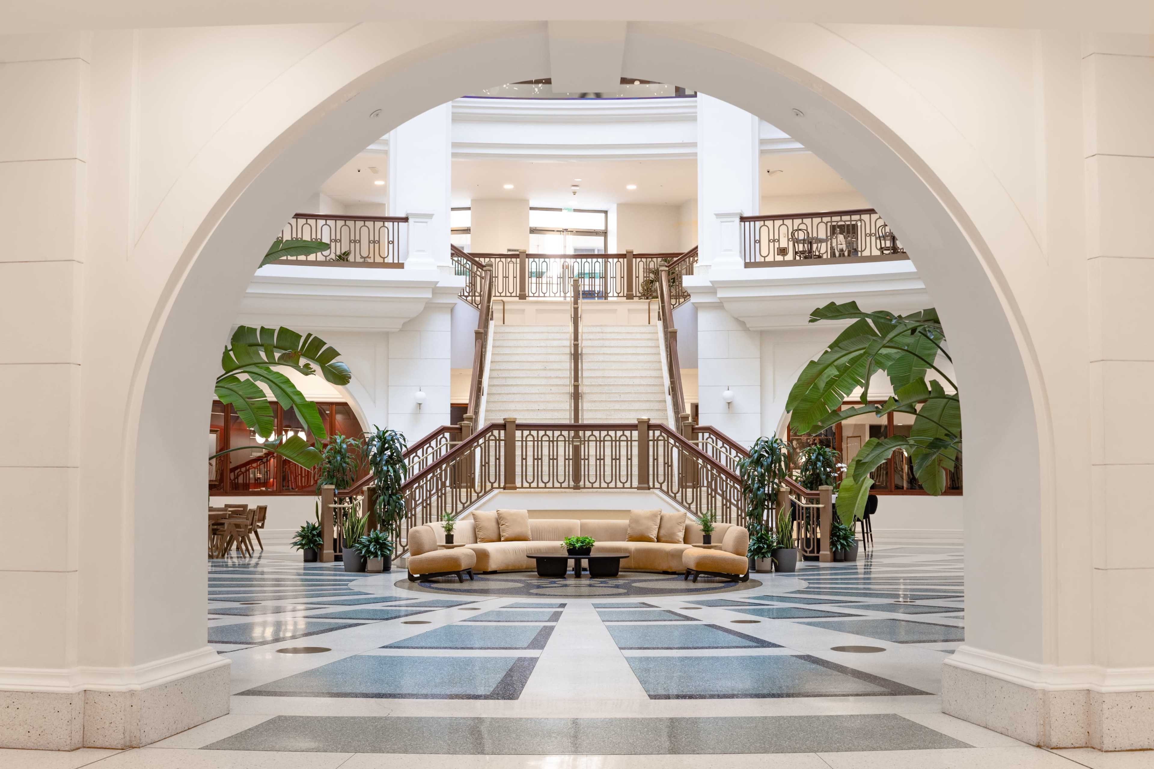 The image shows an interior view of a spacious atrium with a curved archway, featuring a staircase, a circular seating area, and large indoor plants.