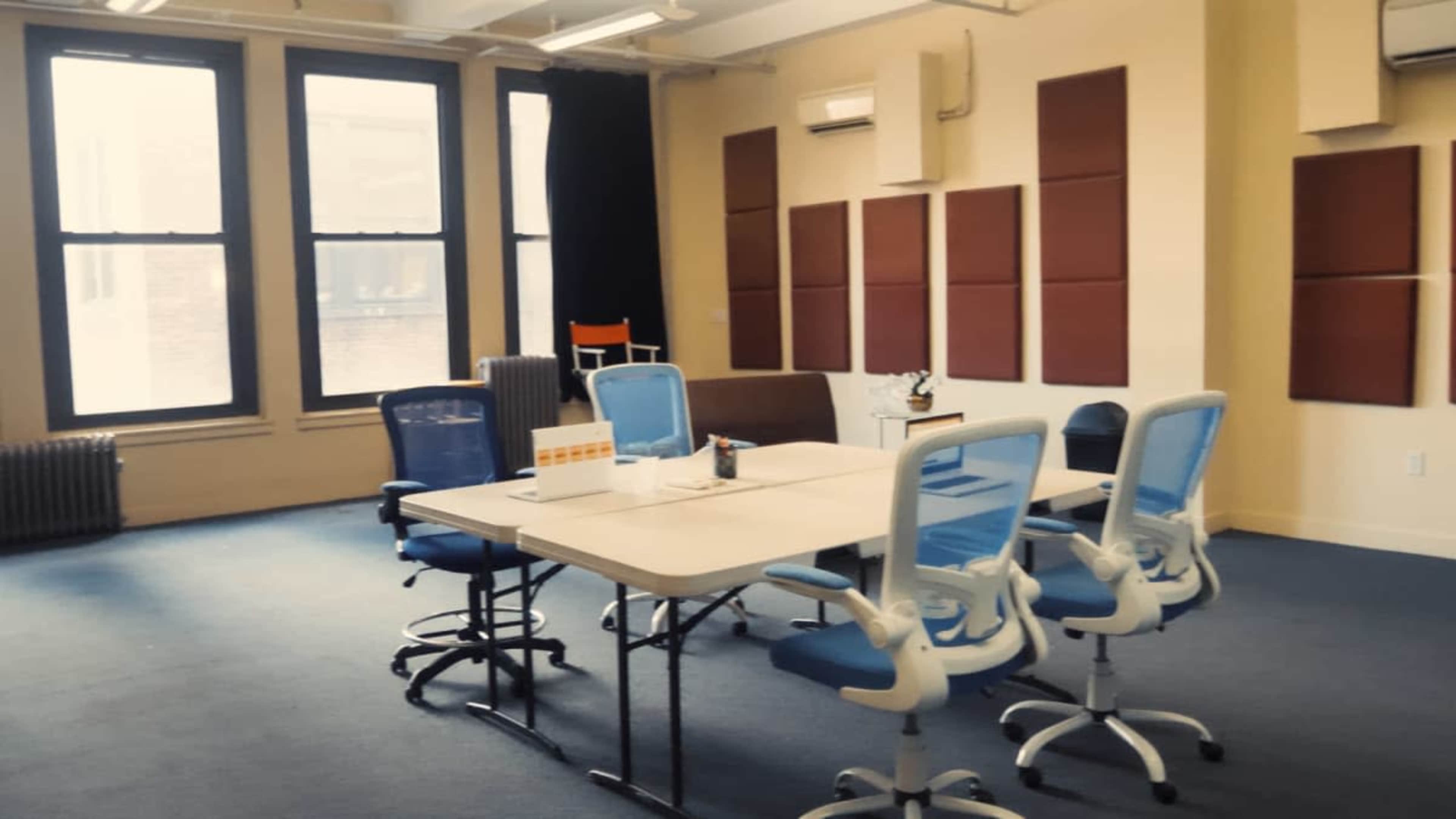 The image shows a bright, spacious meeting room with a large table surrounded by blue chairs and several windows allowing natural light to enter.