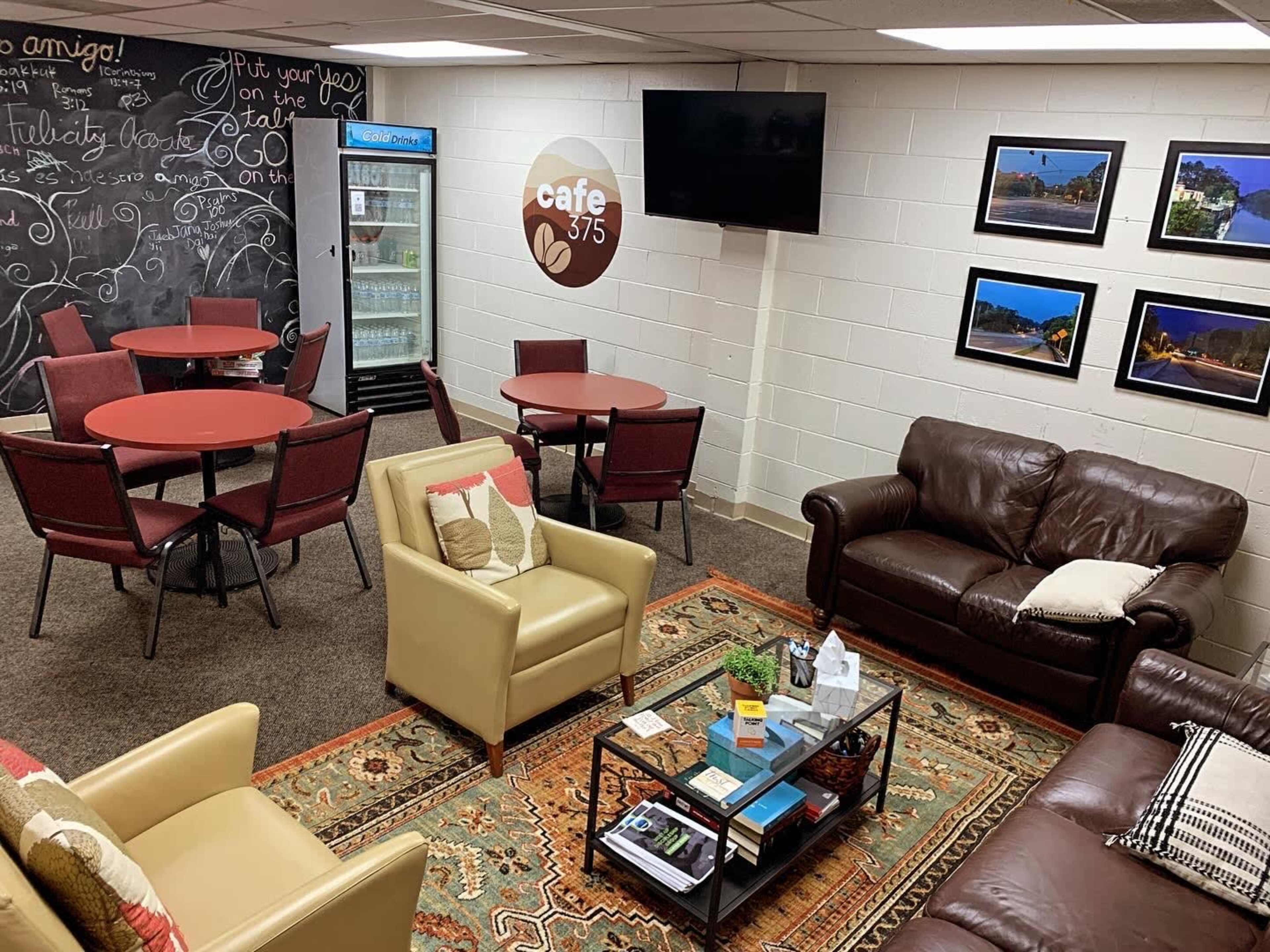 The image shows a cozy lounge area with seating, including two sofas and several round tables, along with a refrigerator and a television on the wall.