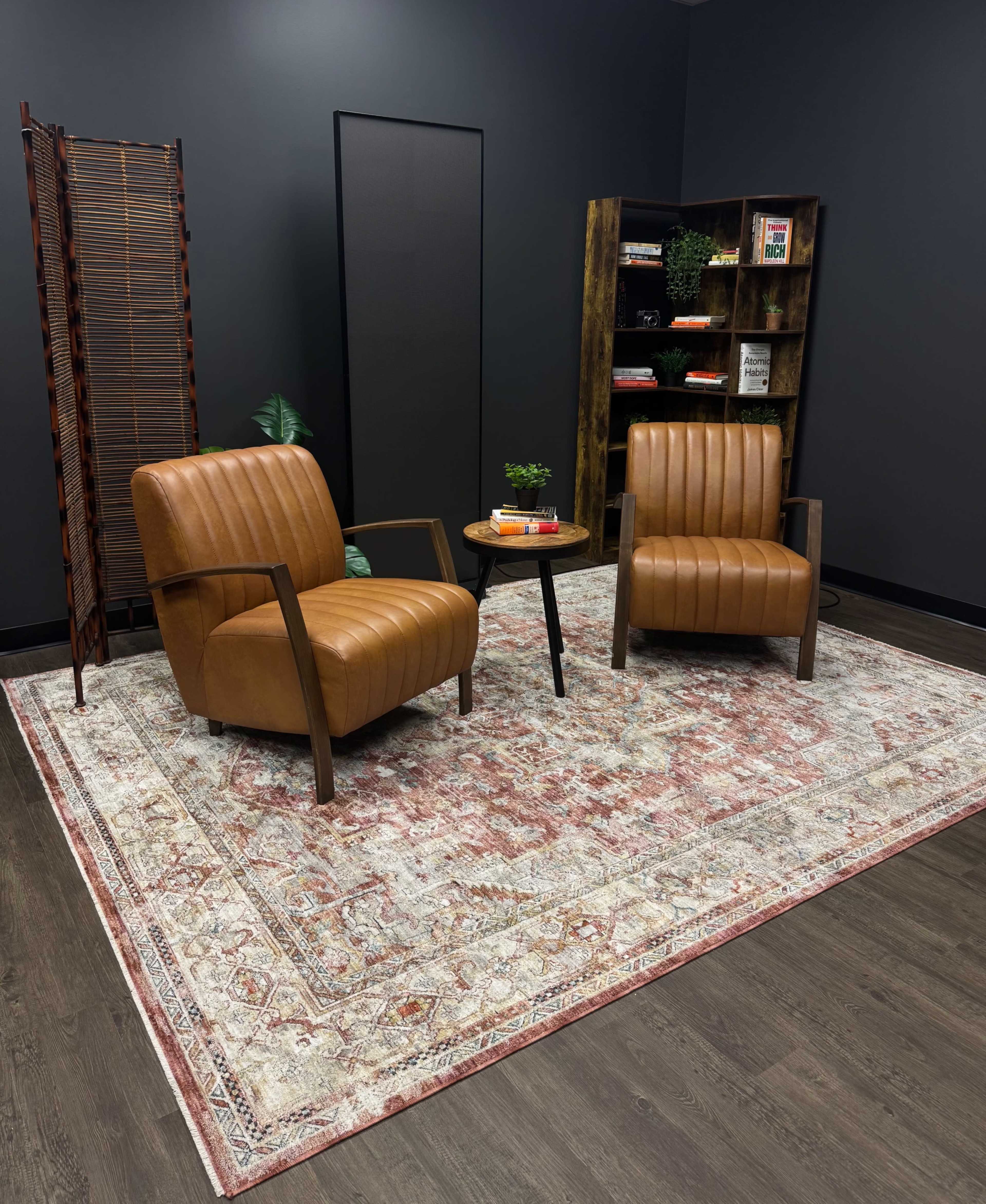 The image shows two brown leather armchairs positioned on a patterned rug in a room with dark walls, a small round table between them, and a bookshelf in the background.