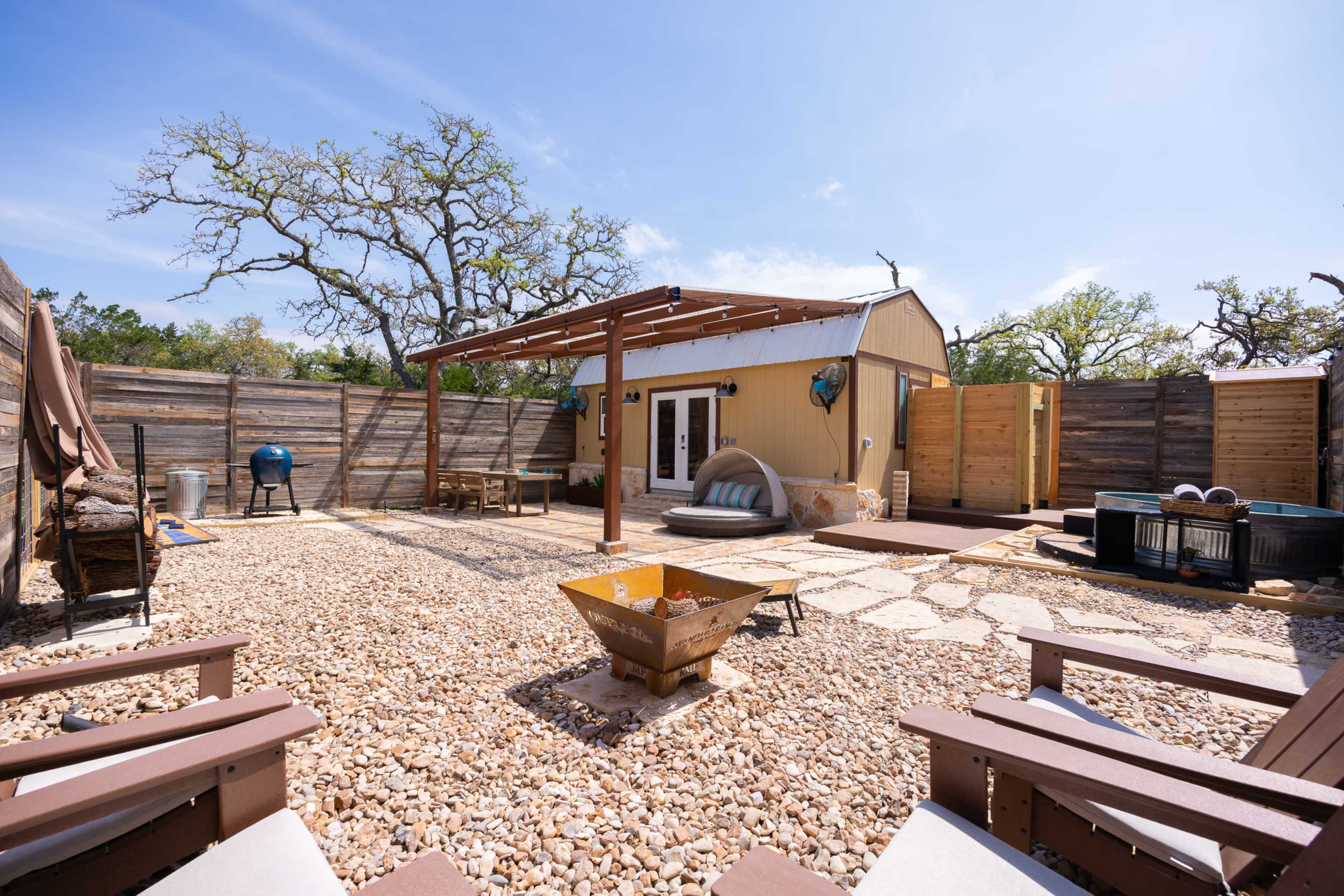 The image shows a landscaped outdoor space with a small structure, seating areas, and a fire pit surrounded by gravel and wooden fencing.