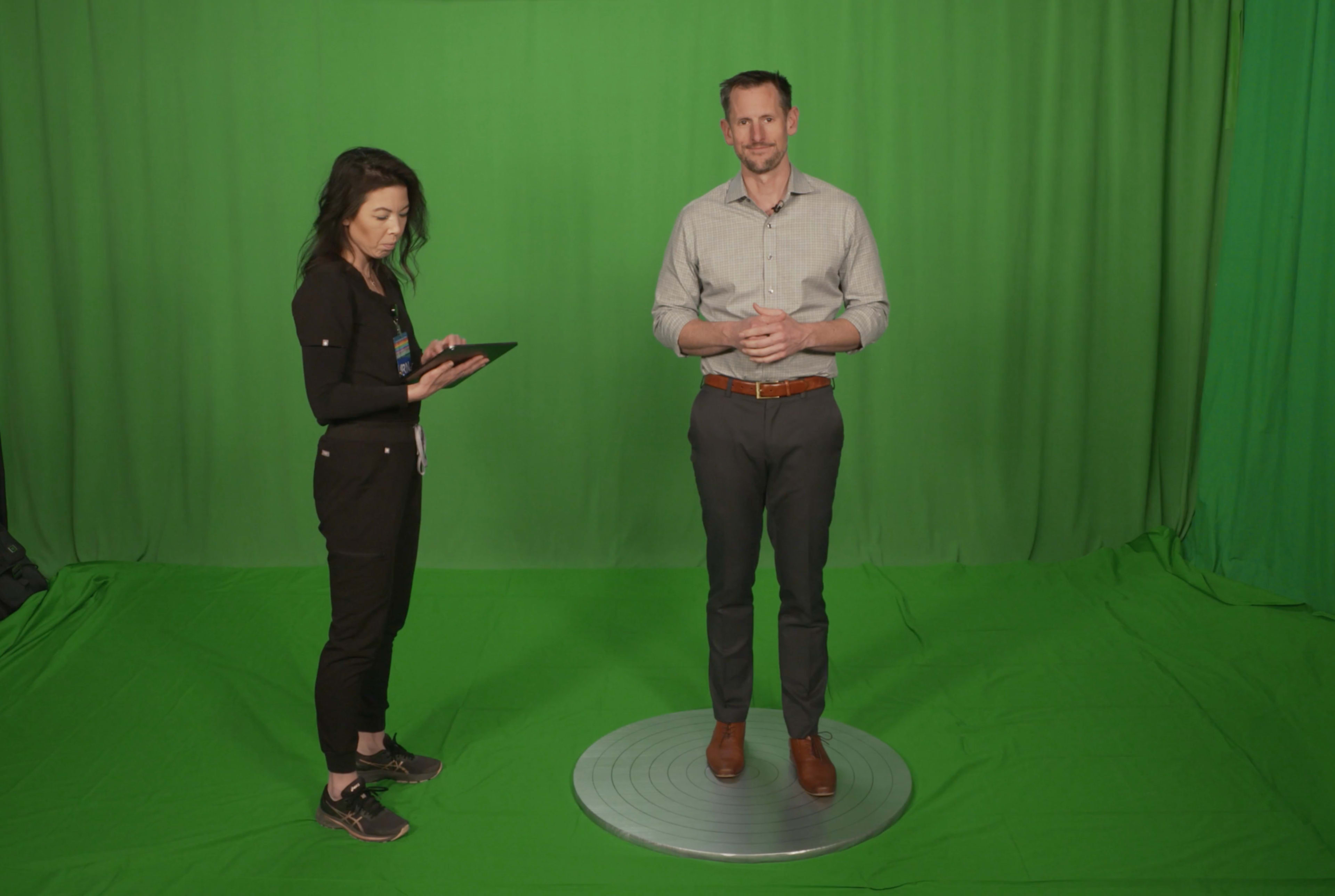 A man stands on a circular platform in front of a green screen while a woman uses a tablet nearby.