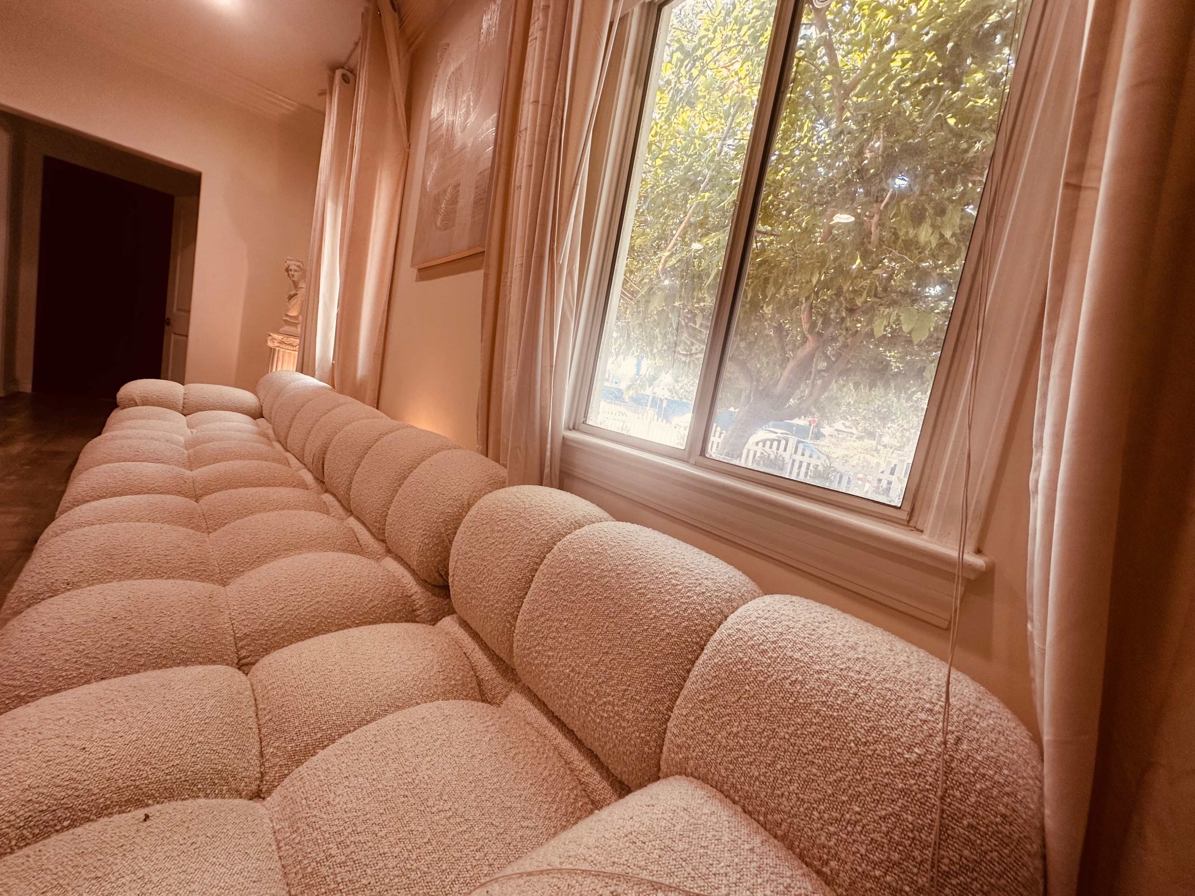 A light-colored, cushioned sofa lines the wall beside a large window with partially drawn curtains, allowing sunlight to filter in.