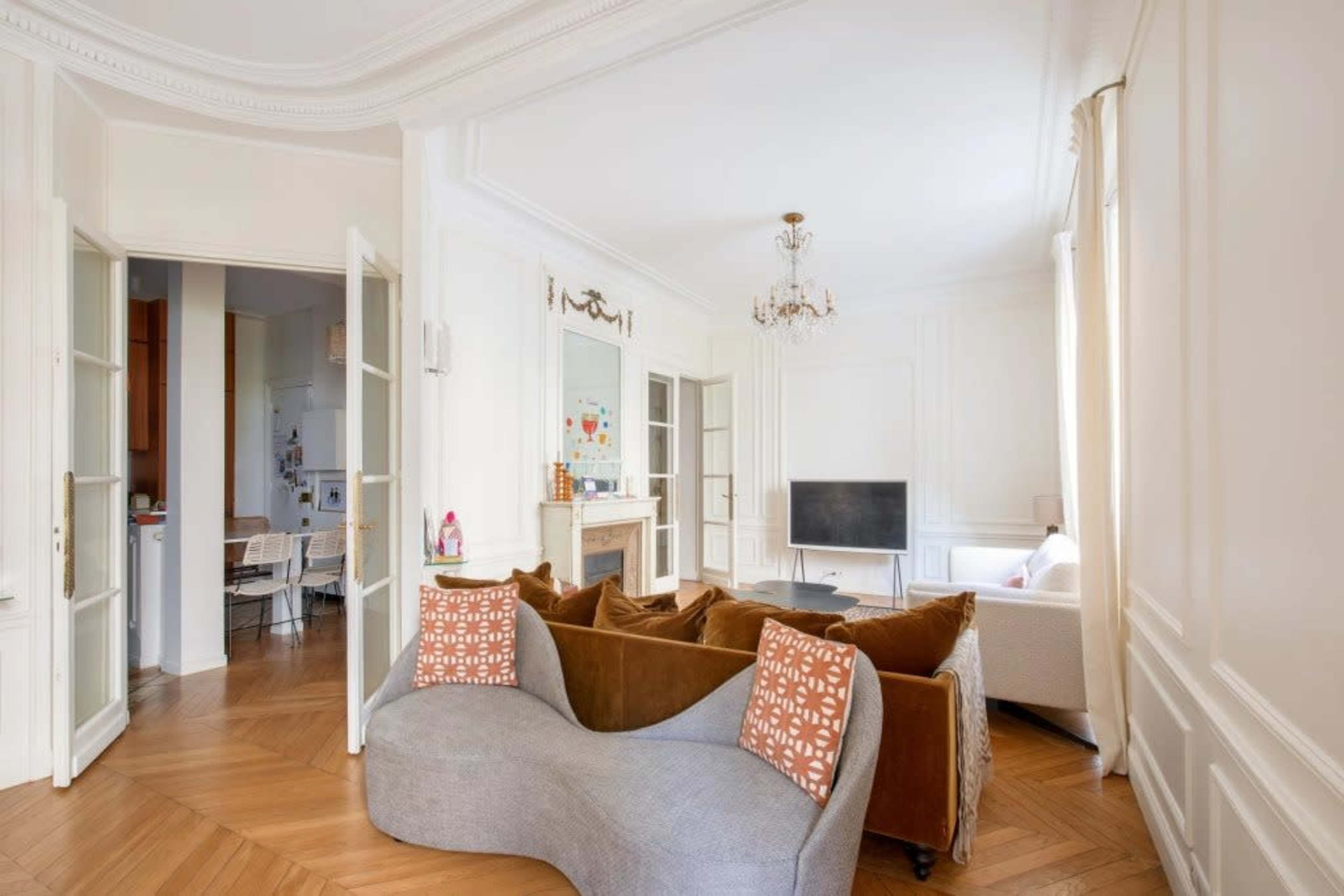 The image features a stylish living room with a unique curved brown sofa, a television, and a dining area visible through an open doorway.