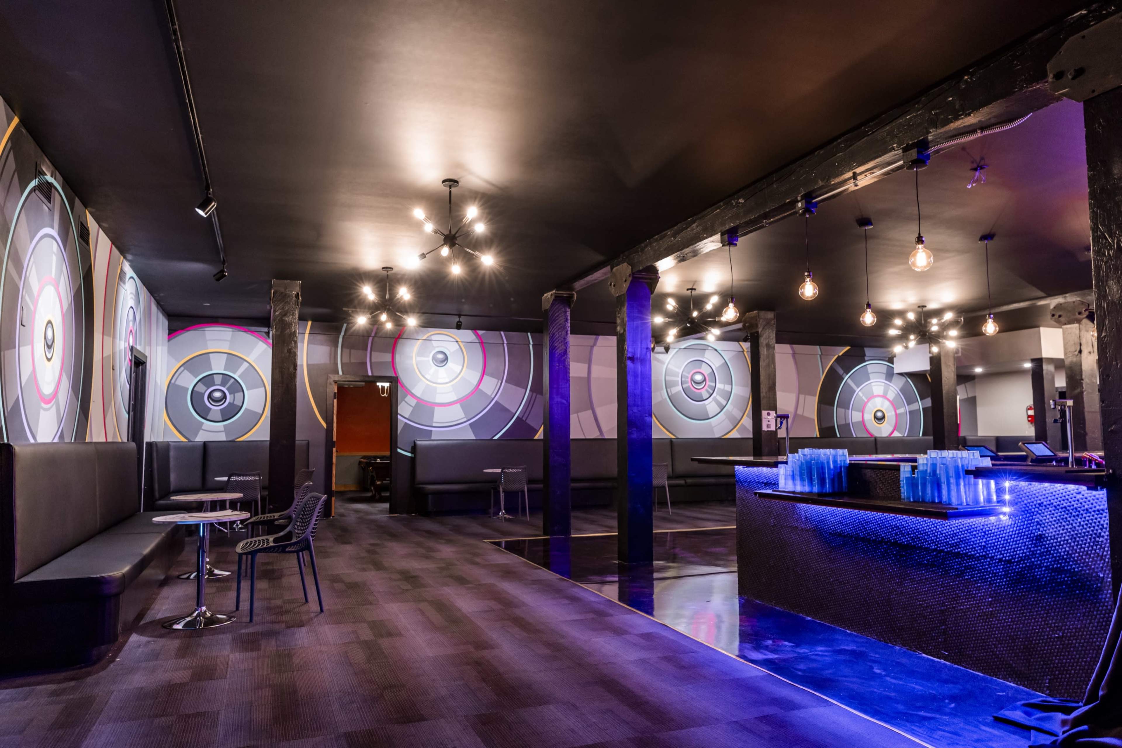 The image shows a modern dart bar interior with colorful target murals on the walls, seating areas along the sides, and a blue-lit bar at the back.