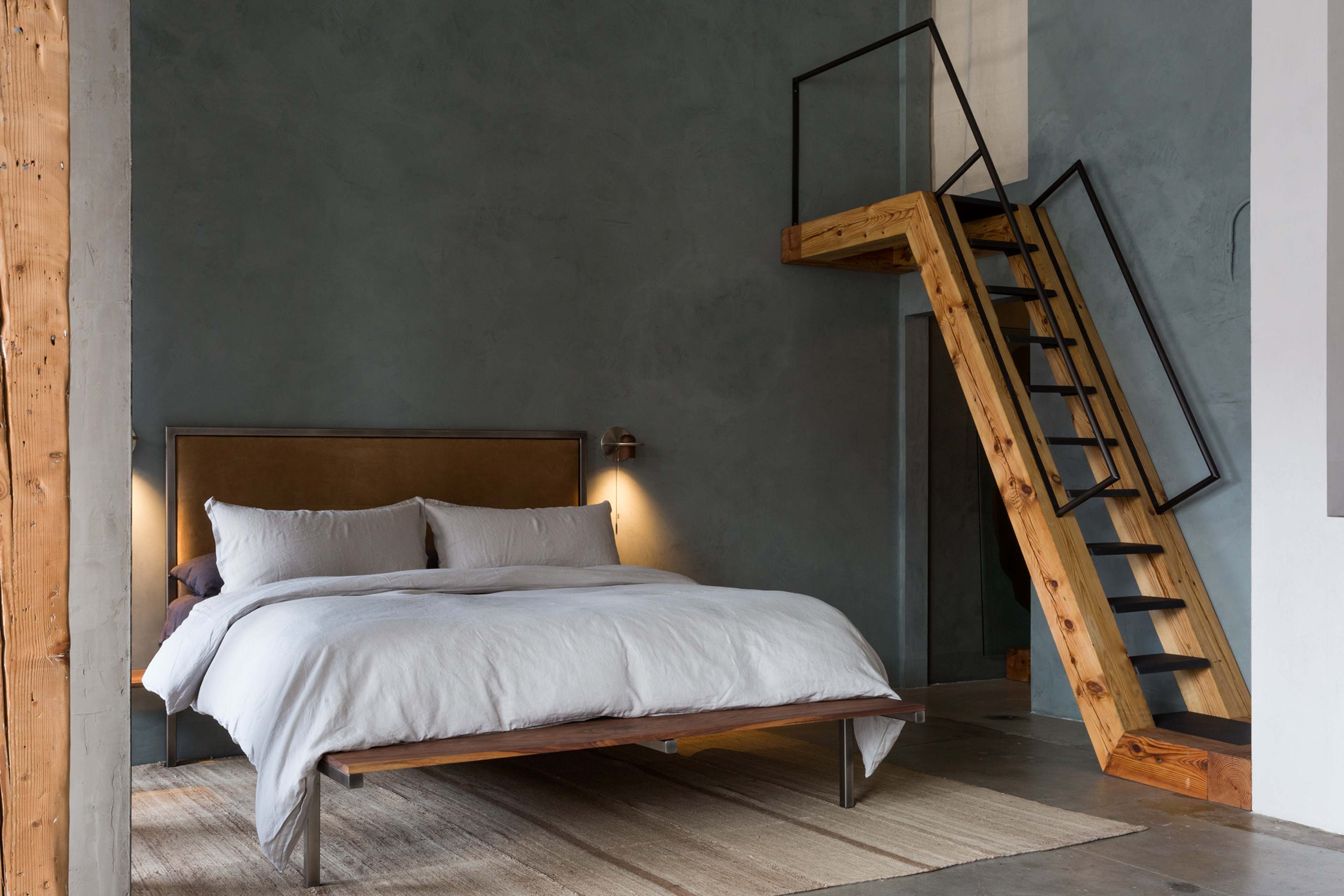 A modern bedroom features a large bed with gray bedding, a wooden headboard, and a staircase made of wood and metal leading to a loft area.
