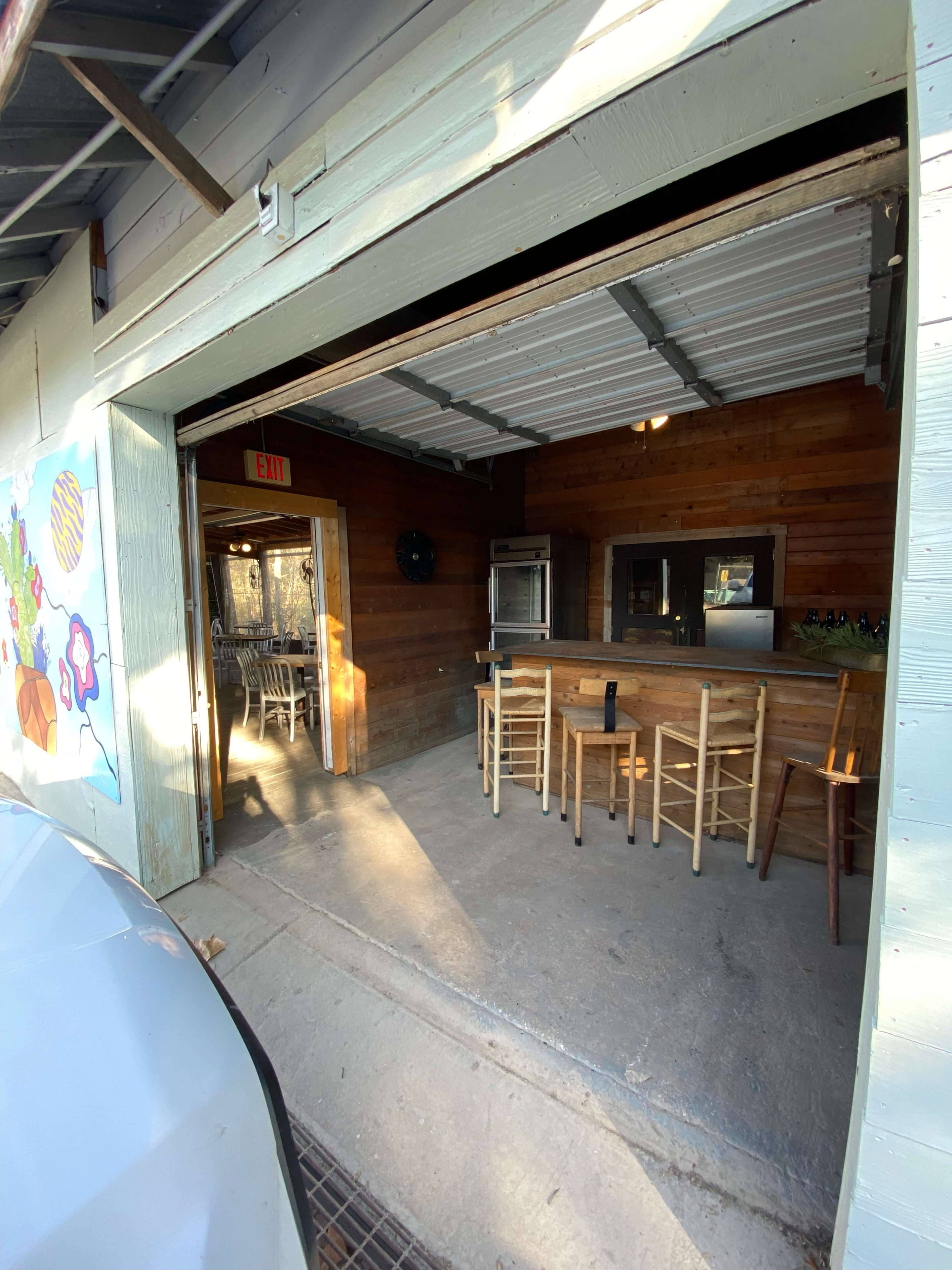 An open garage-like space with a wooden bar and several stools, featuring light streaming in through the entrance.
