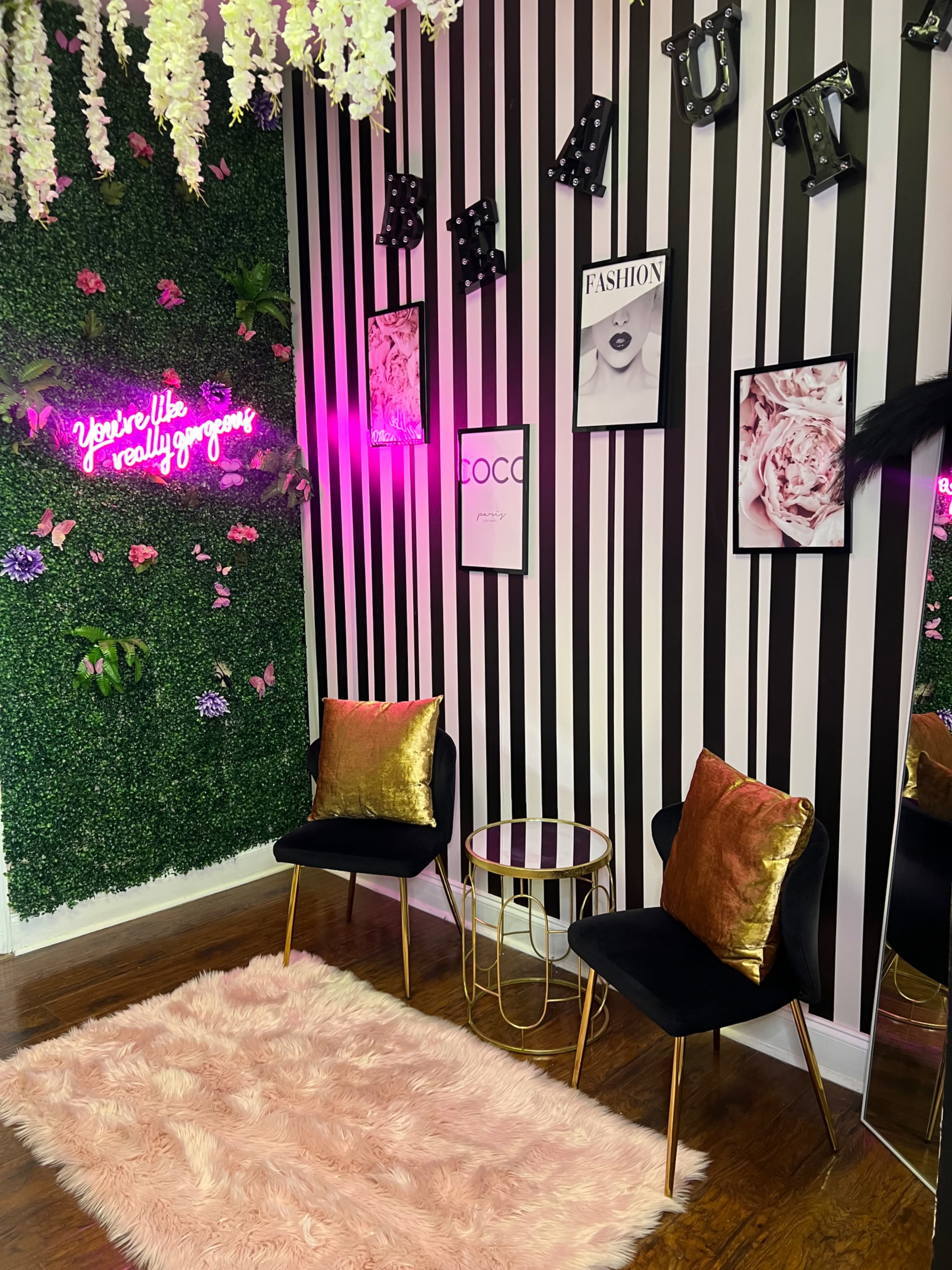 The image shows a stylish room with black and white striped walls, two black chairs with gold accents, a pink fuzzy rug, and various decorative items, including framed pictures and neon signs.