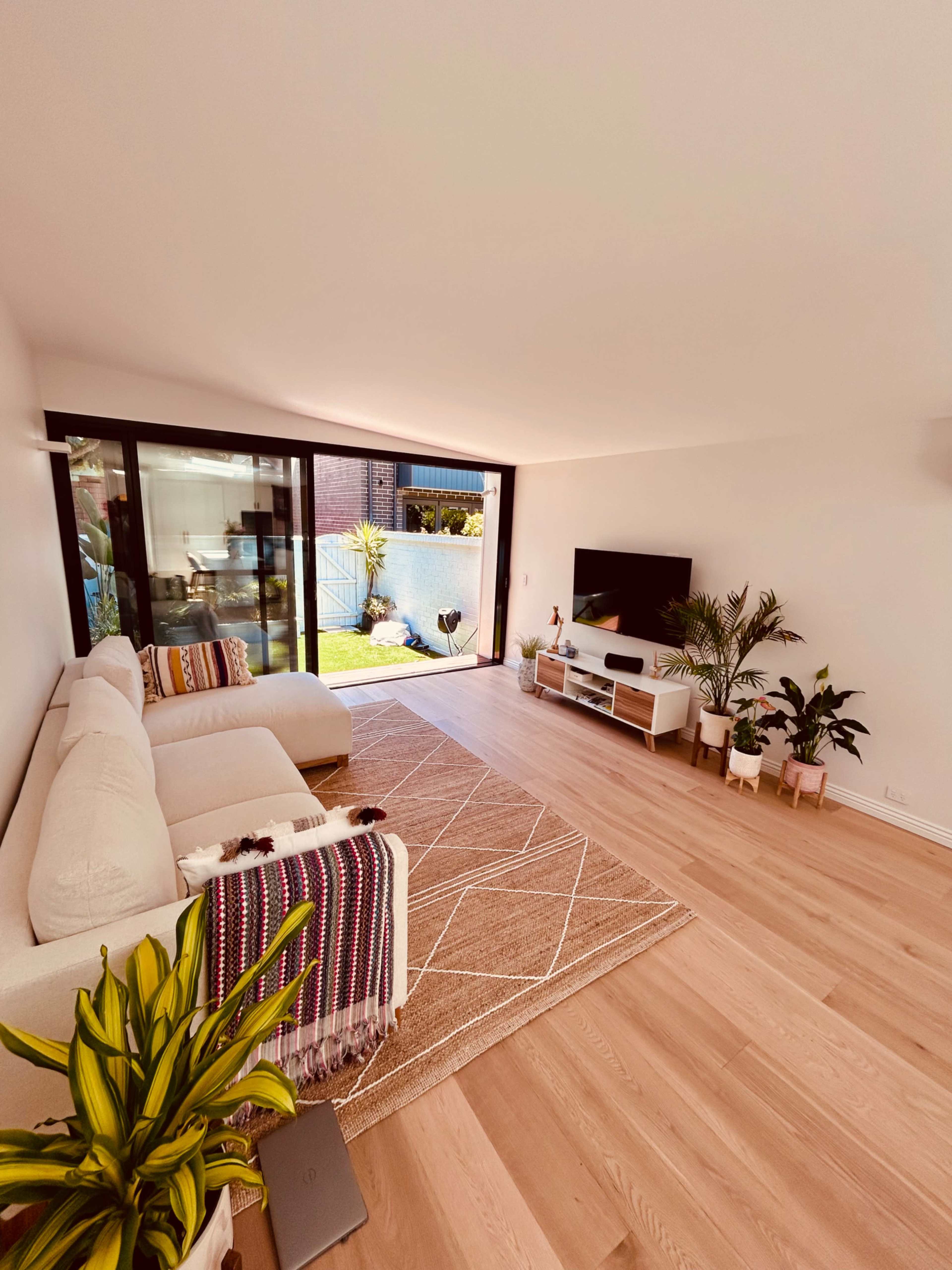 A modern living room features a light-colored section sofa, a large area rug, and a television mounted on the wall, with large windows providing a view of a garden.