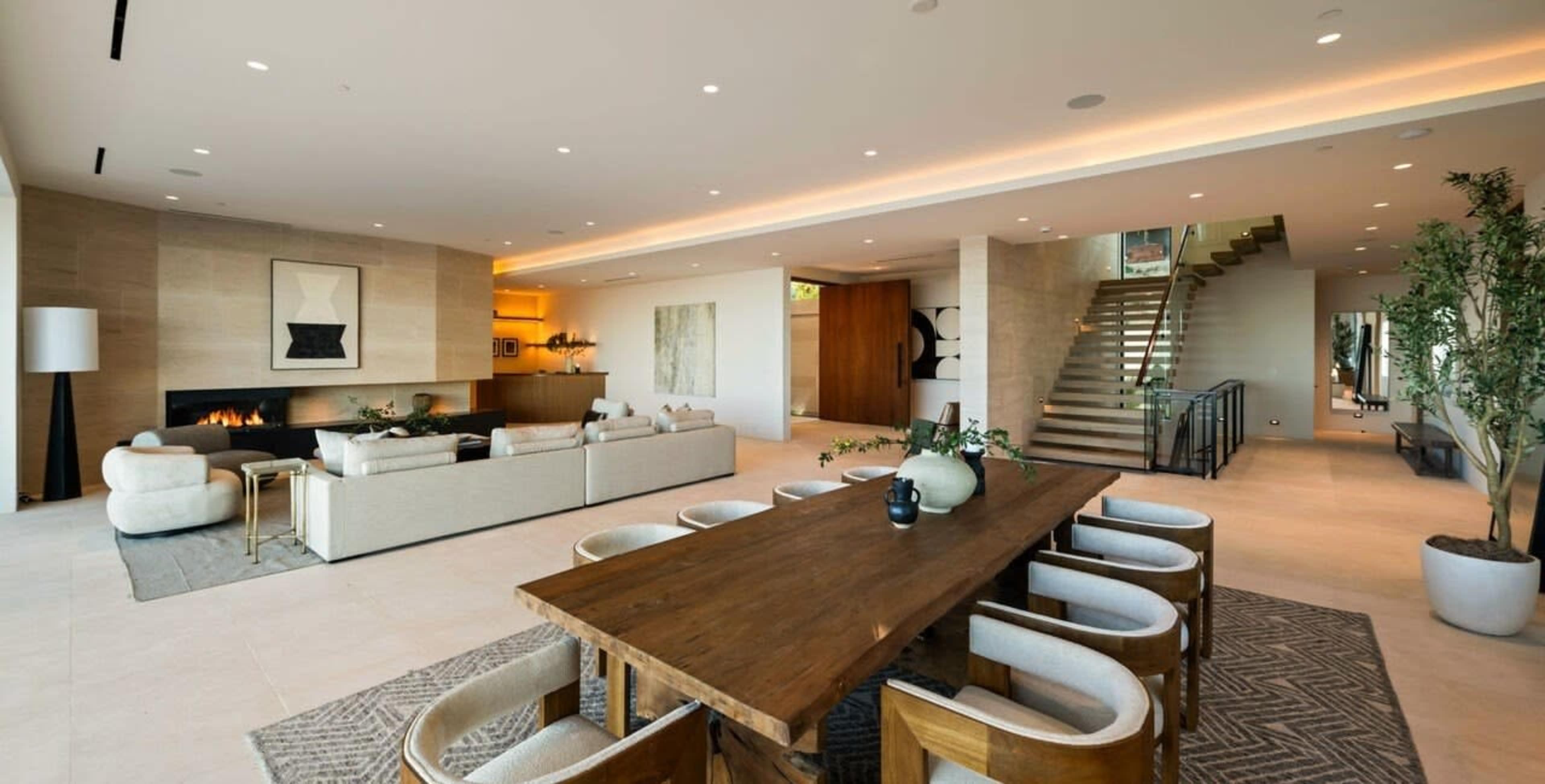 The image shows a spacious, modern living area featuring a dining table, a seating area with sofas, and a staircase leading to an upper level.