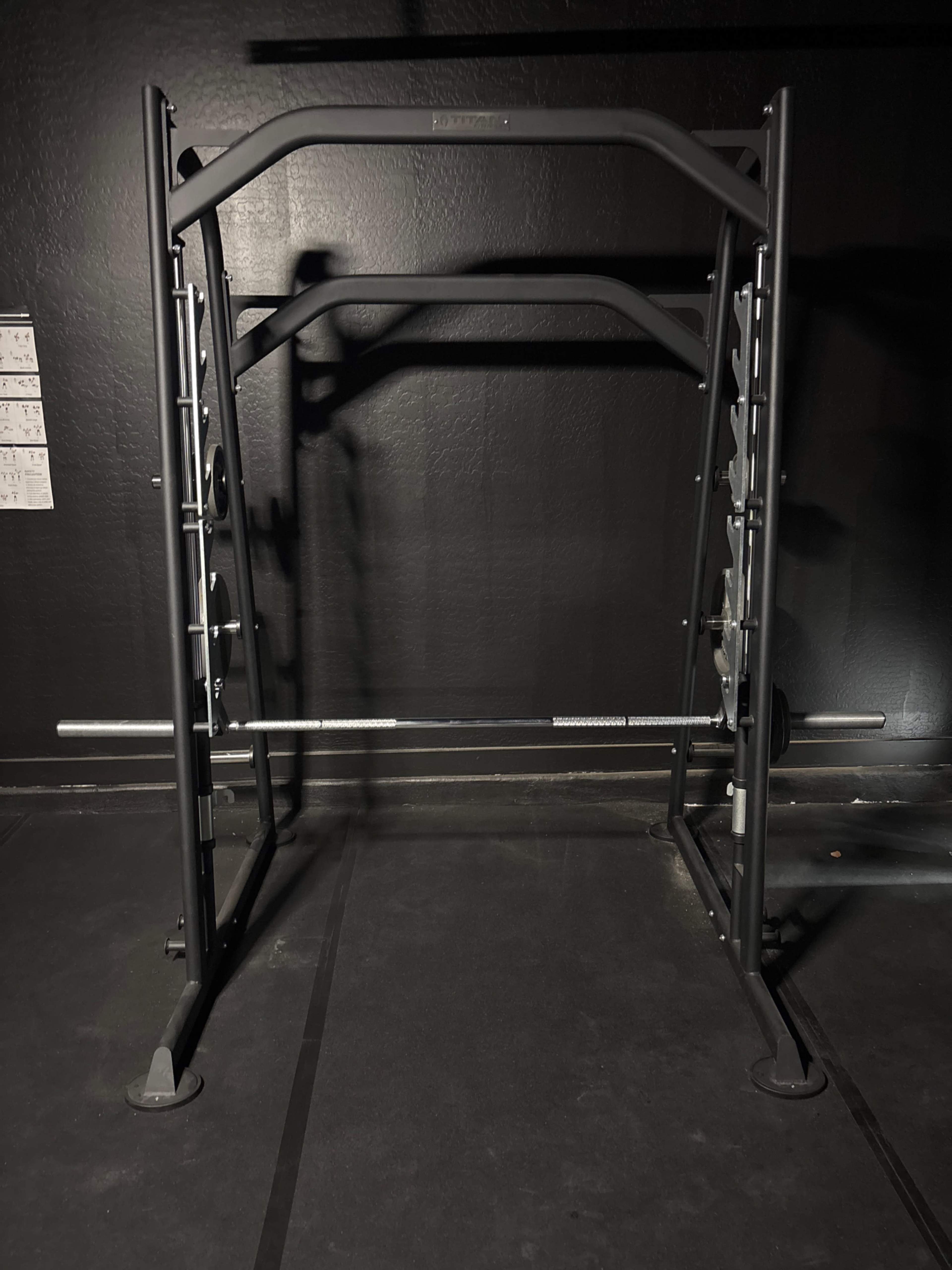 The image shows a squat rack in a gym setting, positioned against a black wall with a barbell placed on its supports.