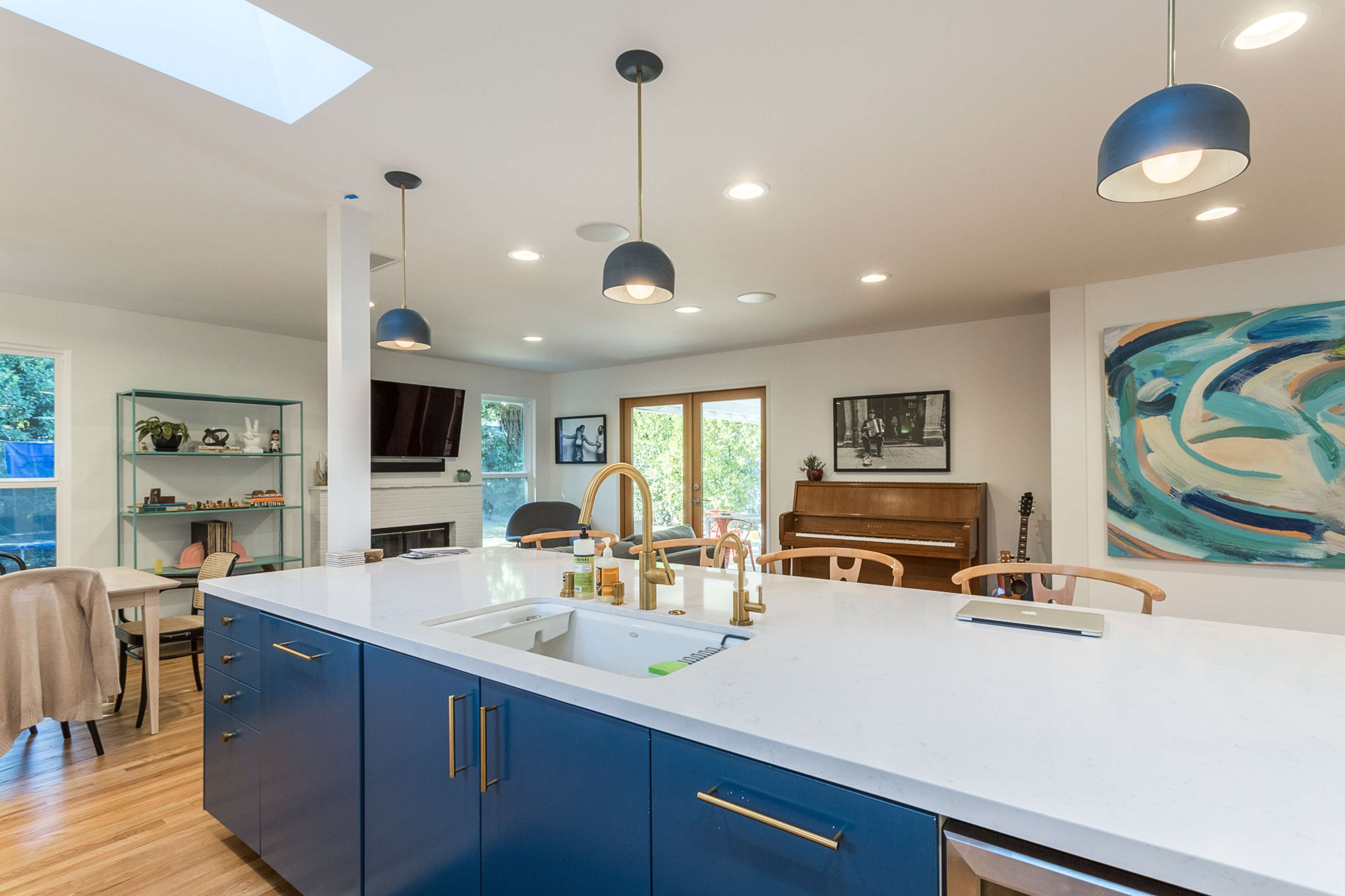 The image shows a modern kitchen with a central island, blue cabinetry, a white countertop, and a dining area featuring a piano and large windows.