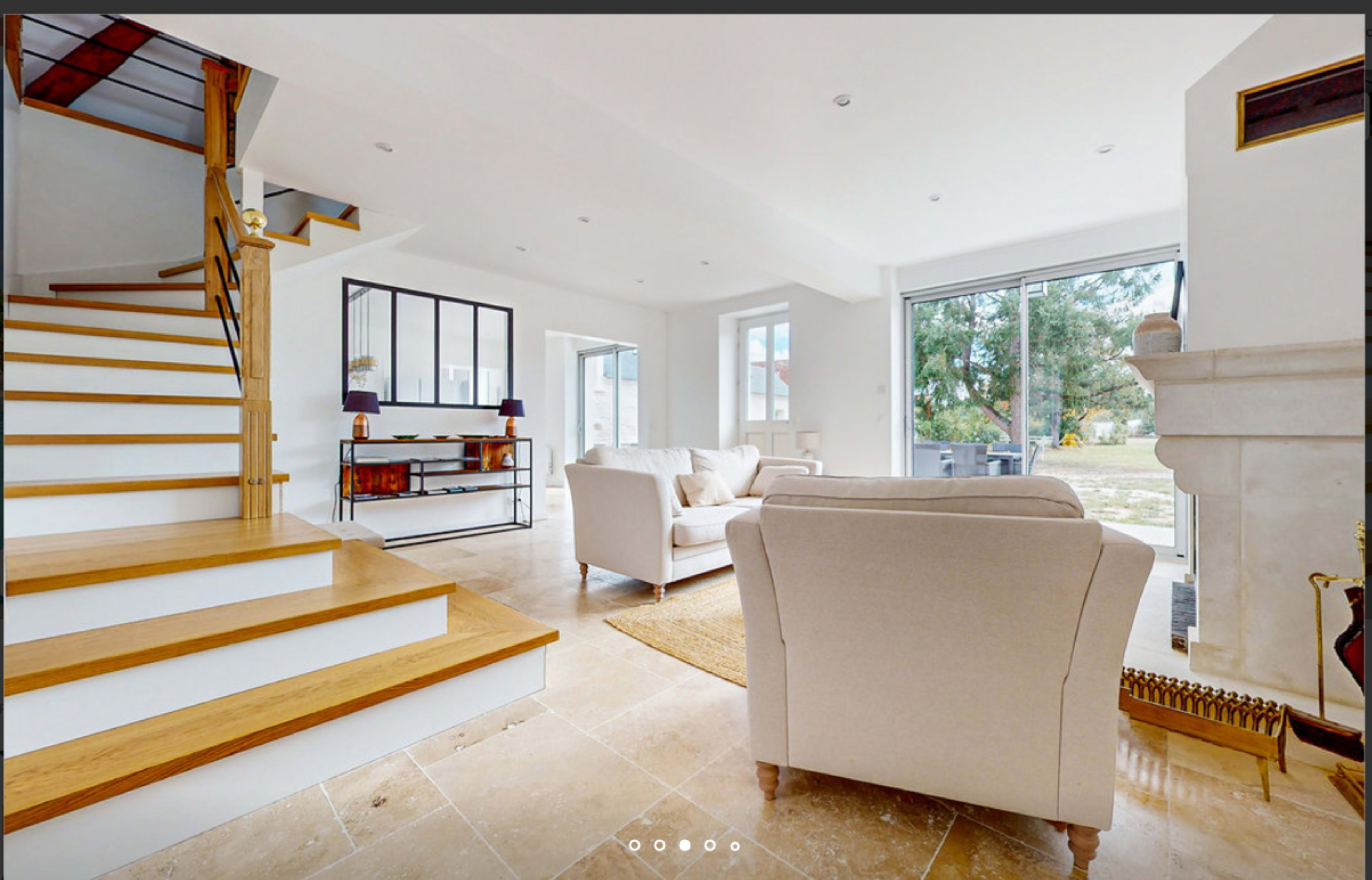 The image shows a bright, open living space with a staircase on the left, a large window in the back, and a beige sofa facing a fireplace.