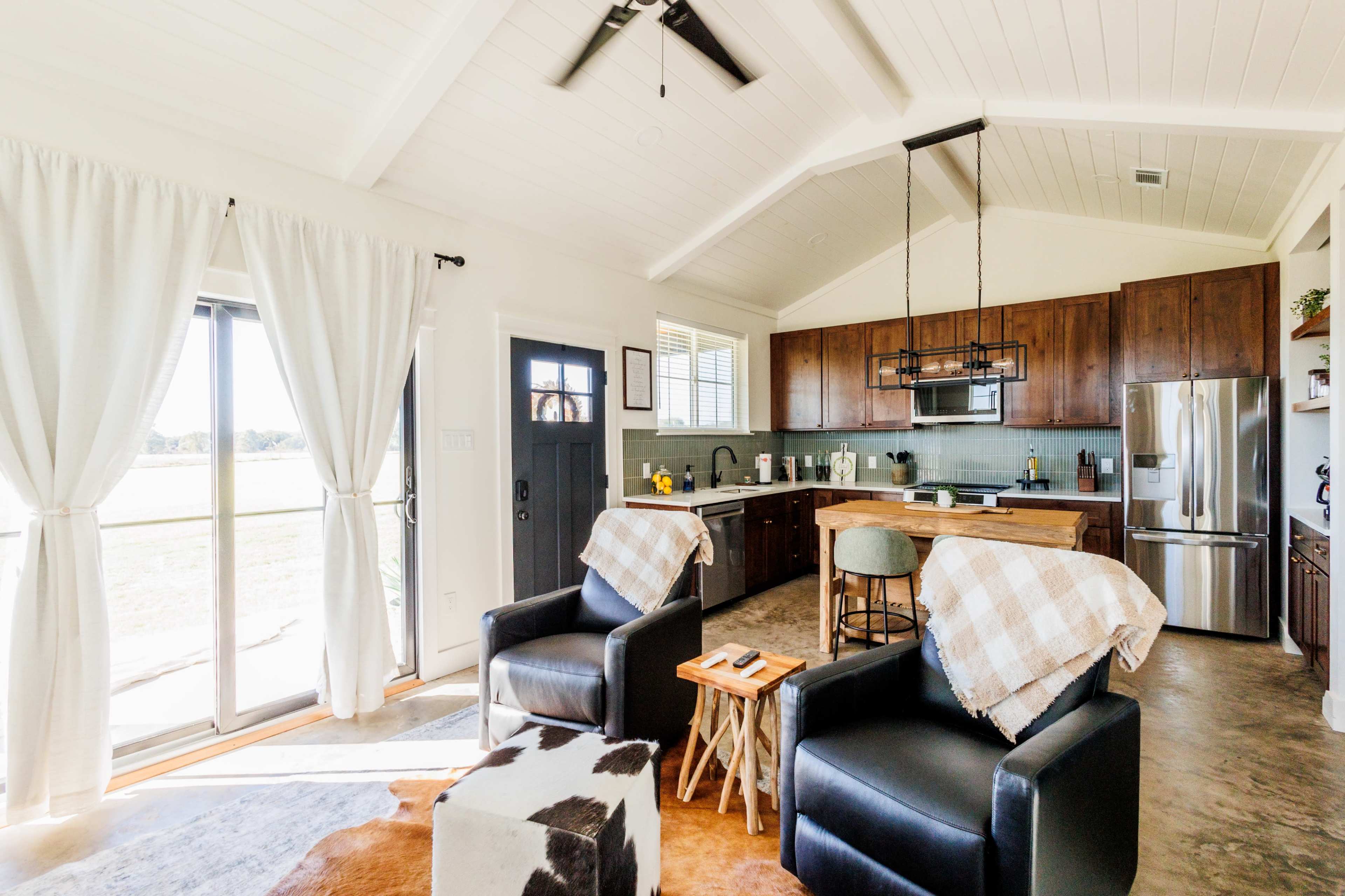 A cozy interior of a modern home features two black chairs, a wooden table, and a well-equipped kitchen with a large window letting in natural light.