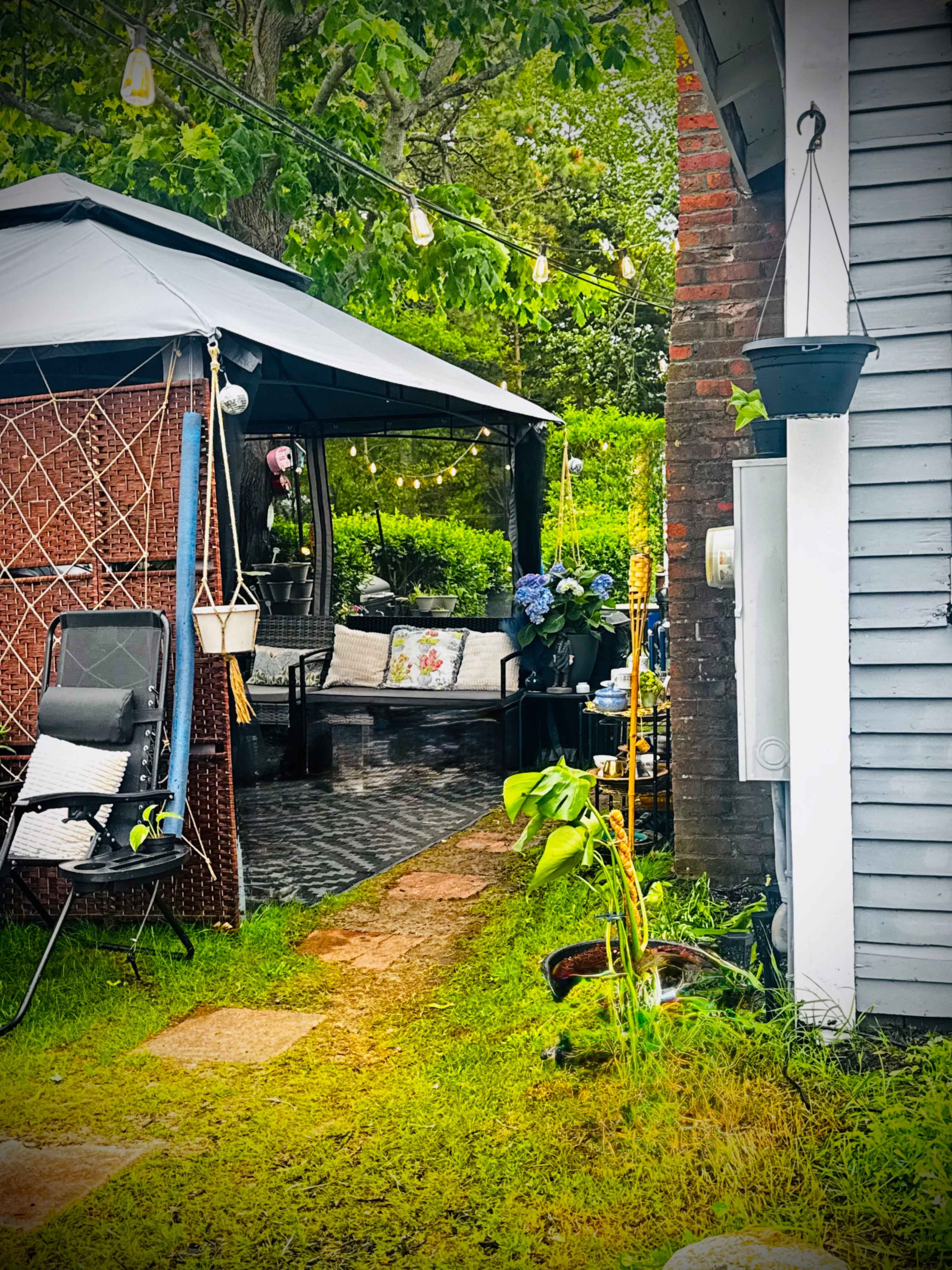 The image features a pathway leading through a garden area with a gazebo, outdoor furniture, and hanging plants beside a wooden structure.