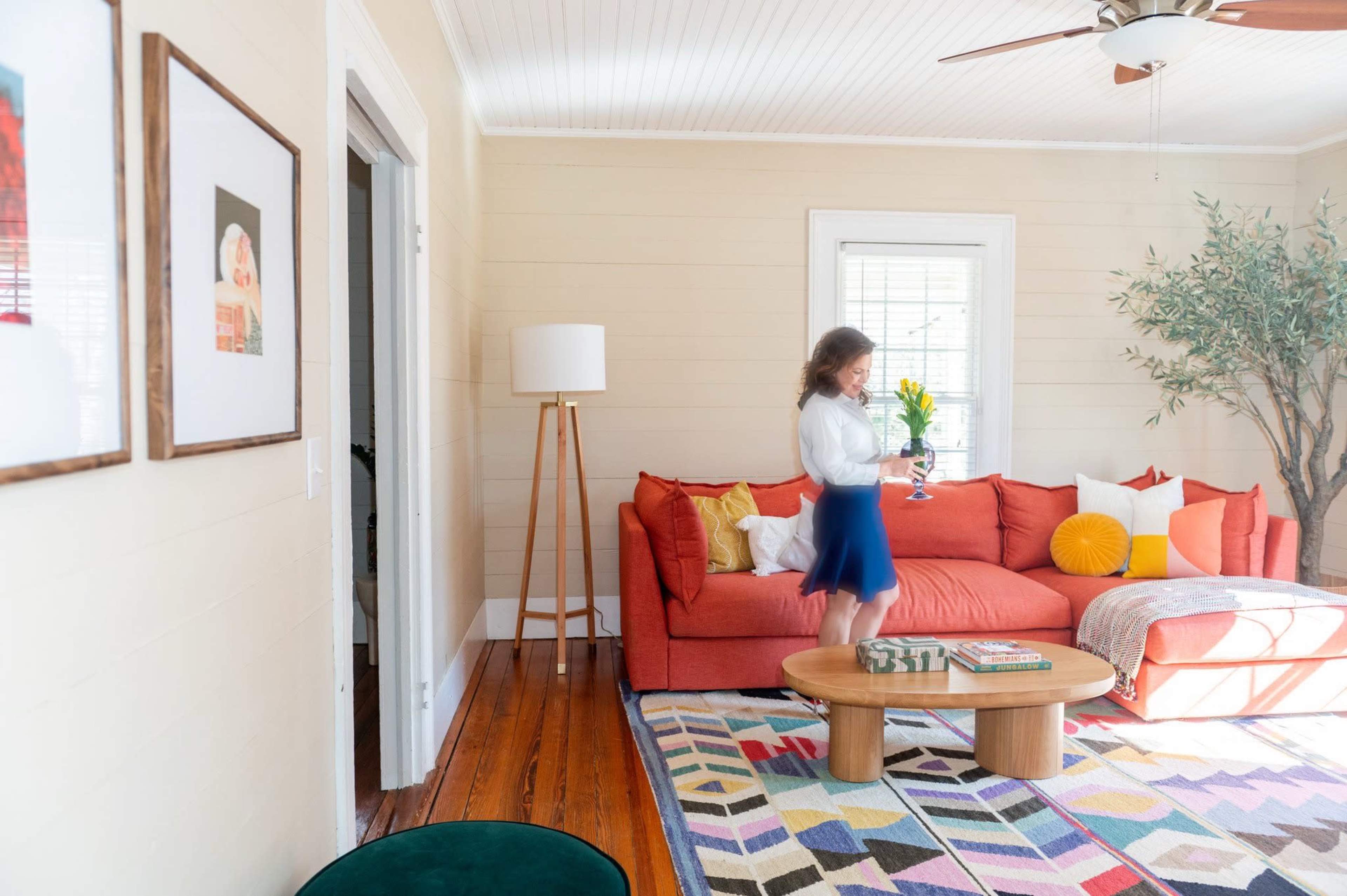 A woman in a white blouse and blue skirt walks through a bright living room featuring a red sofa, a coffee table, and a large area rug.