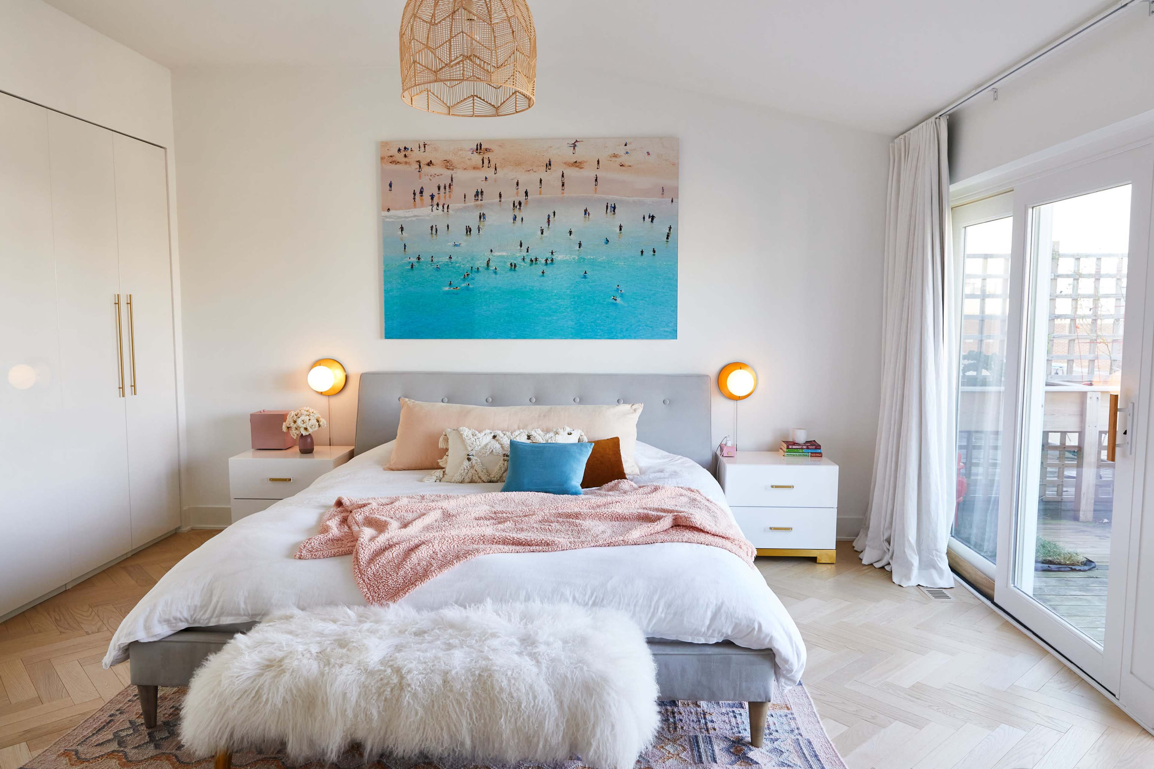 The image shows a bright bedroom featuring a king-size bed with colorful pillows, a large artwork of a beach scene on the wall above, and a sliding glass door leading to an outdoor area.
