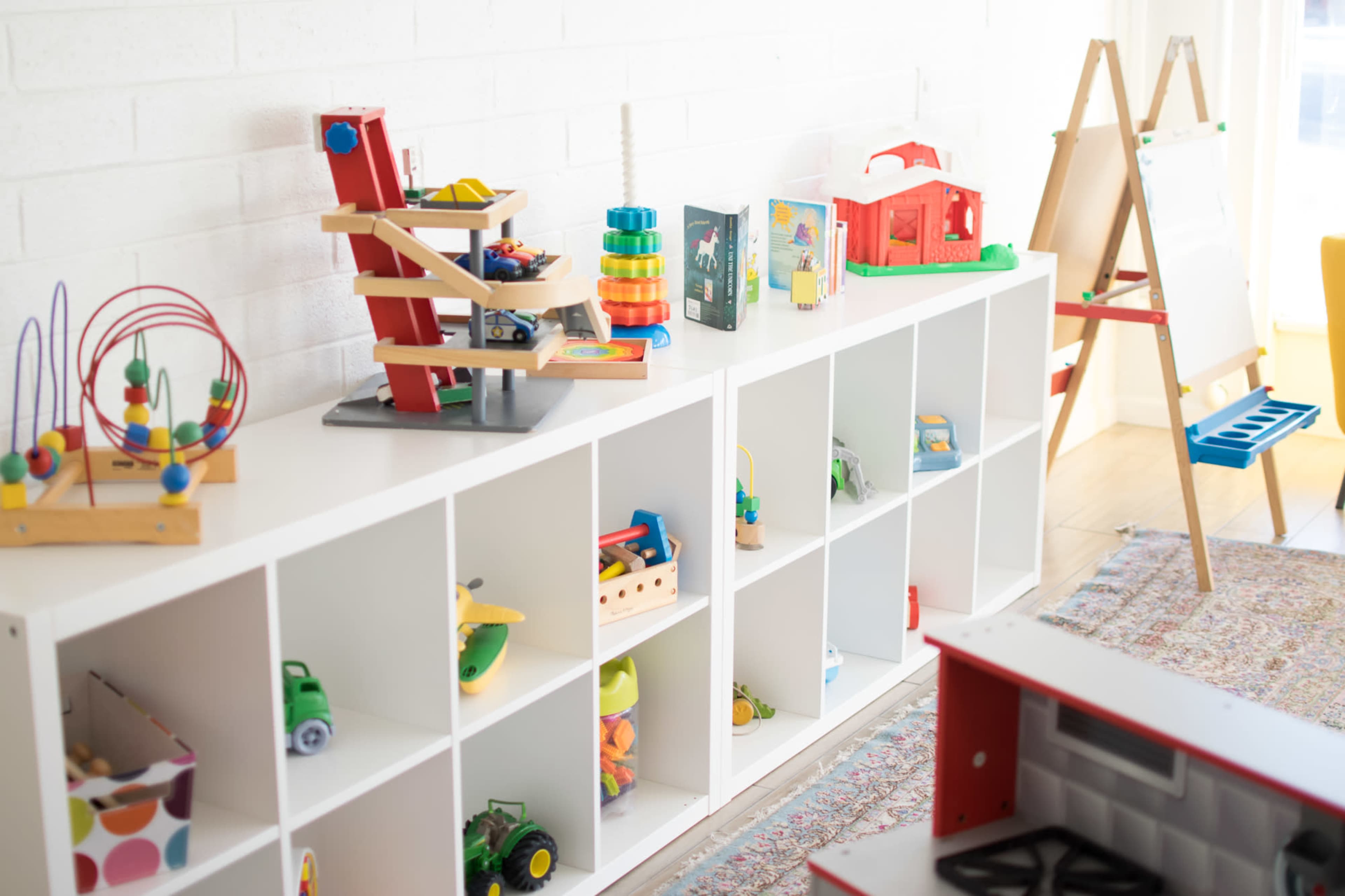 A playroom features a white shelving unit filled with various colorful toys, including stacking rings, a wooden bead maze, and toy vehicles.