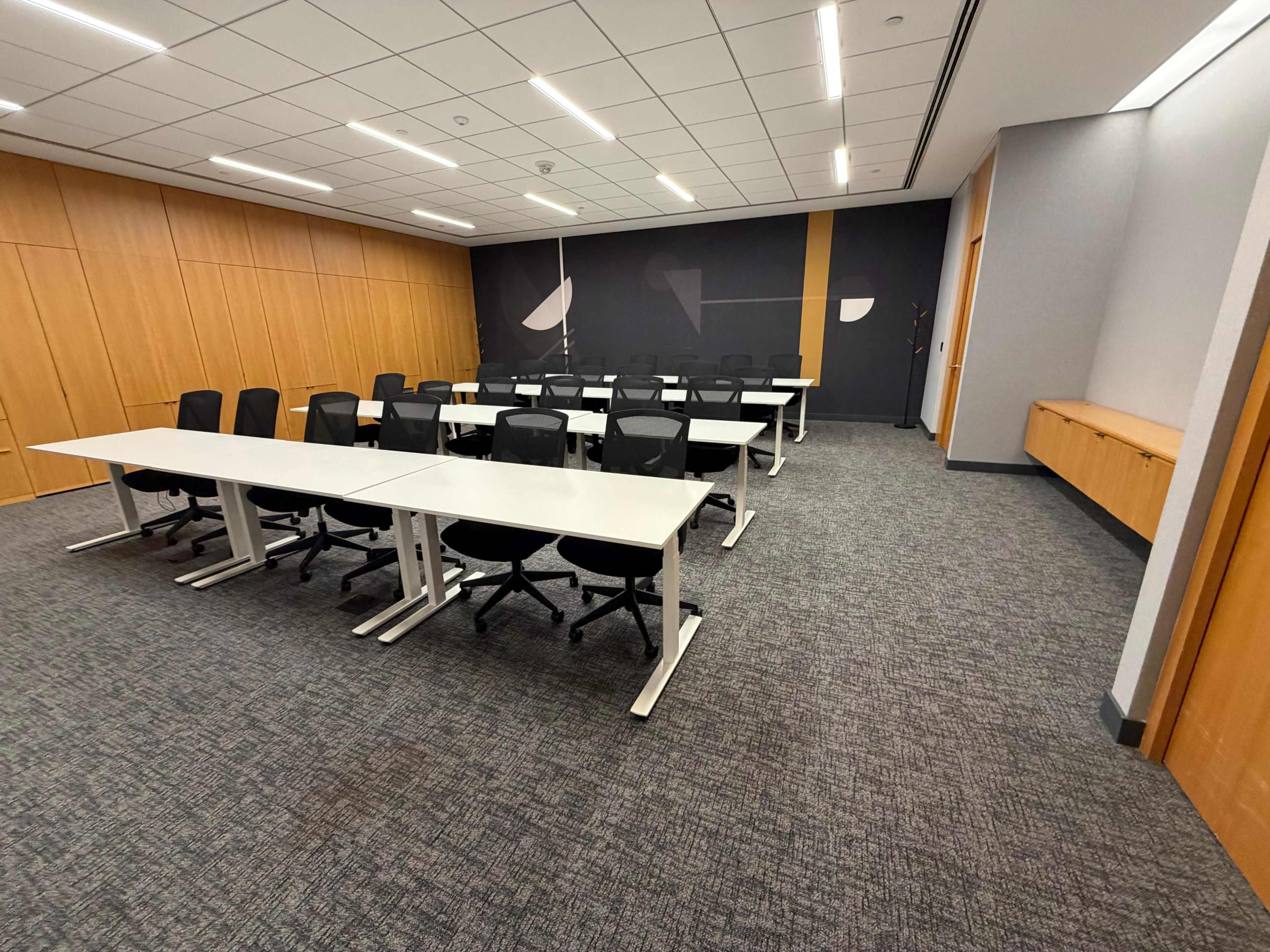 The image shows a modern conference room with several rows of white tables and black chairs arranged neatly on a gray carpet.