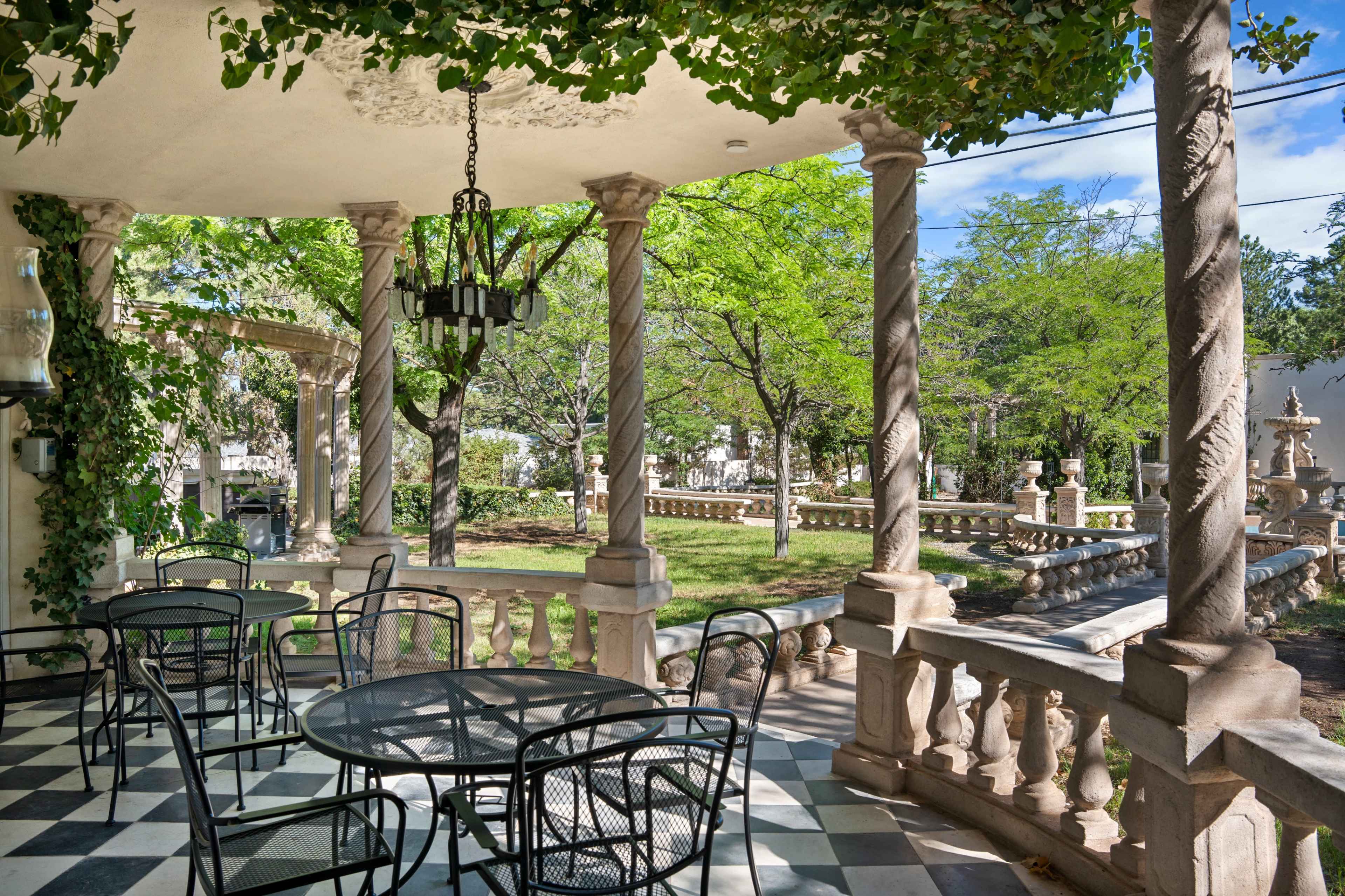 The image shows a garden patio with a black and white checkered floor, adorned with four stone columns, a chandelier, and black metal tables and chairs surrounded by lush greenery.