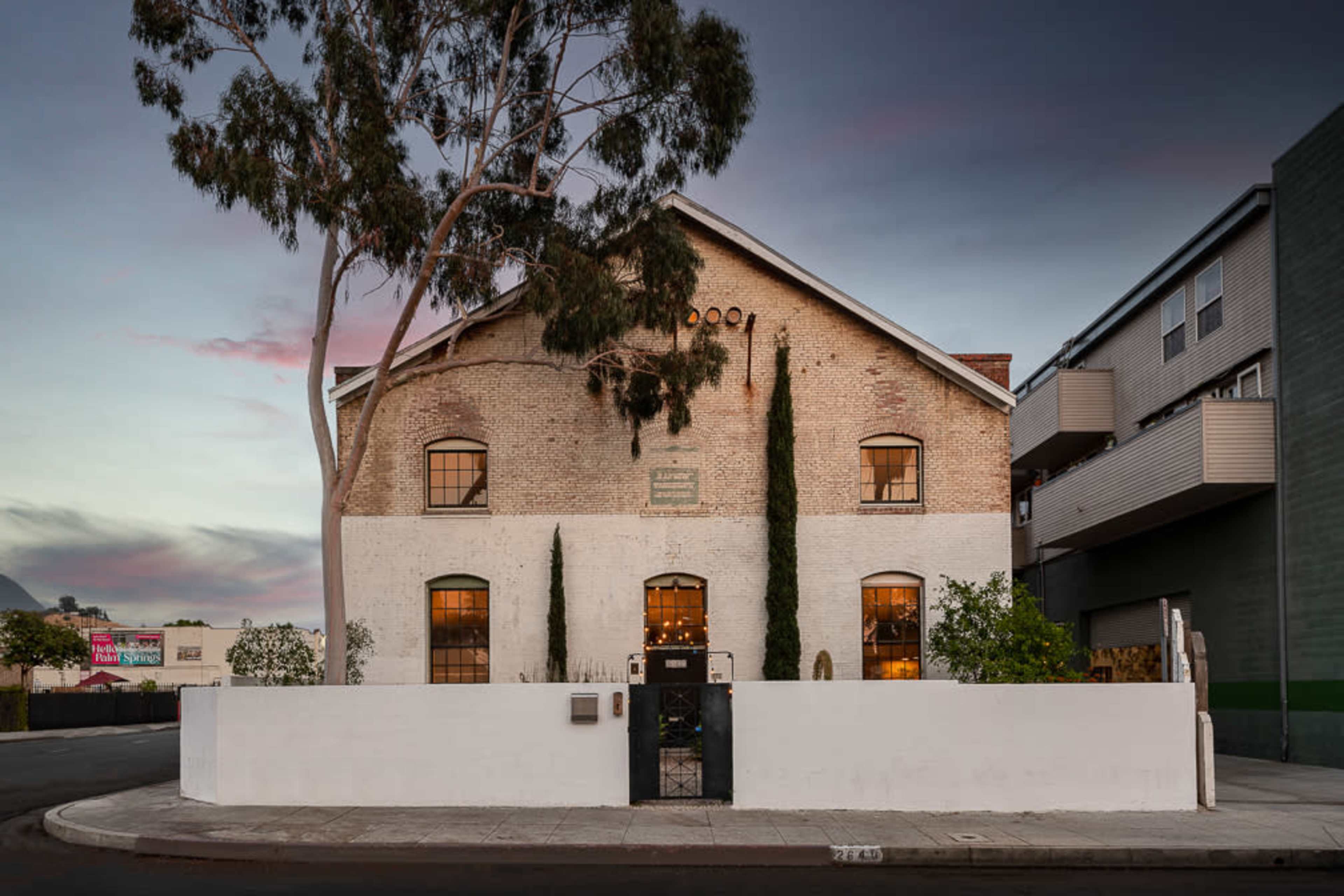 A large, white-walled building with a distinctive, sloped roof, surrounded by trees and other structures on a street corner at dusk.