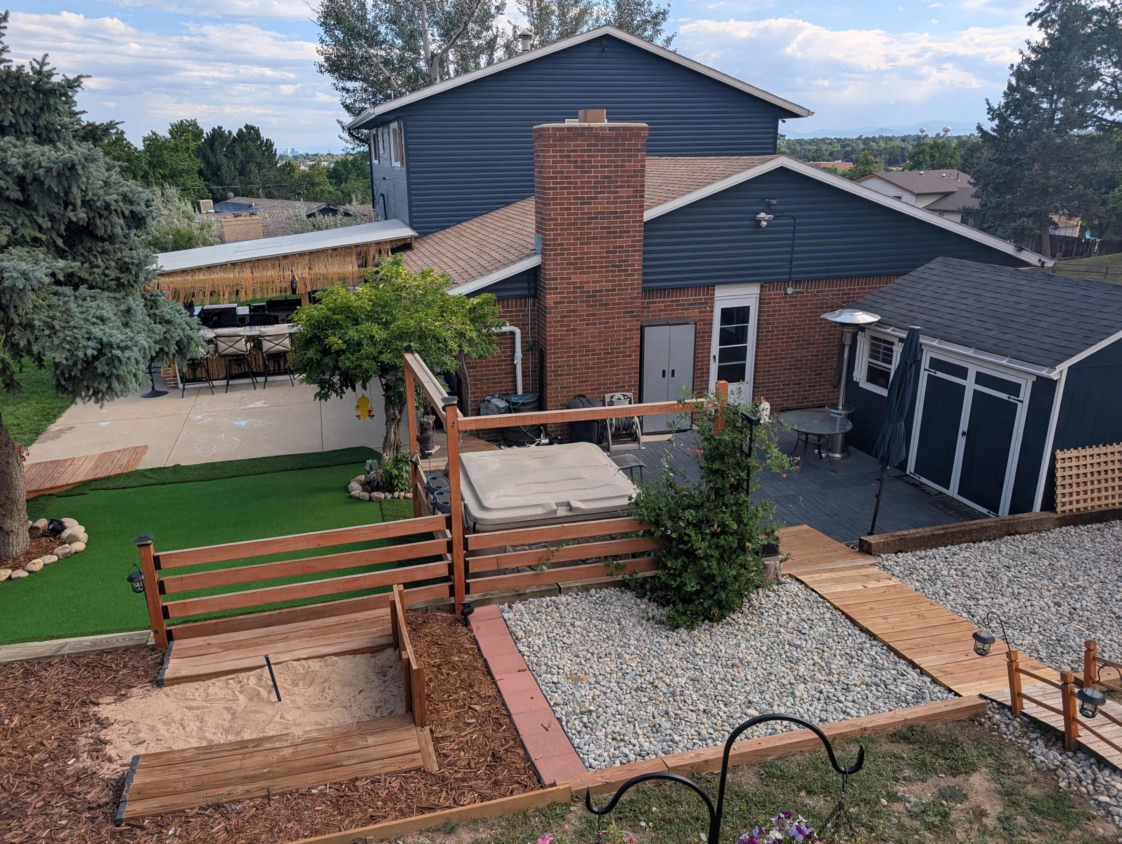 The image shows a two-story house with a mixture of blue siding and brick, surrounded by a landscaped backyard featuring a hot tub, wooden decking, and a sandbox.