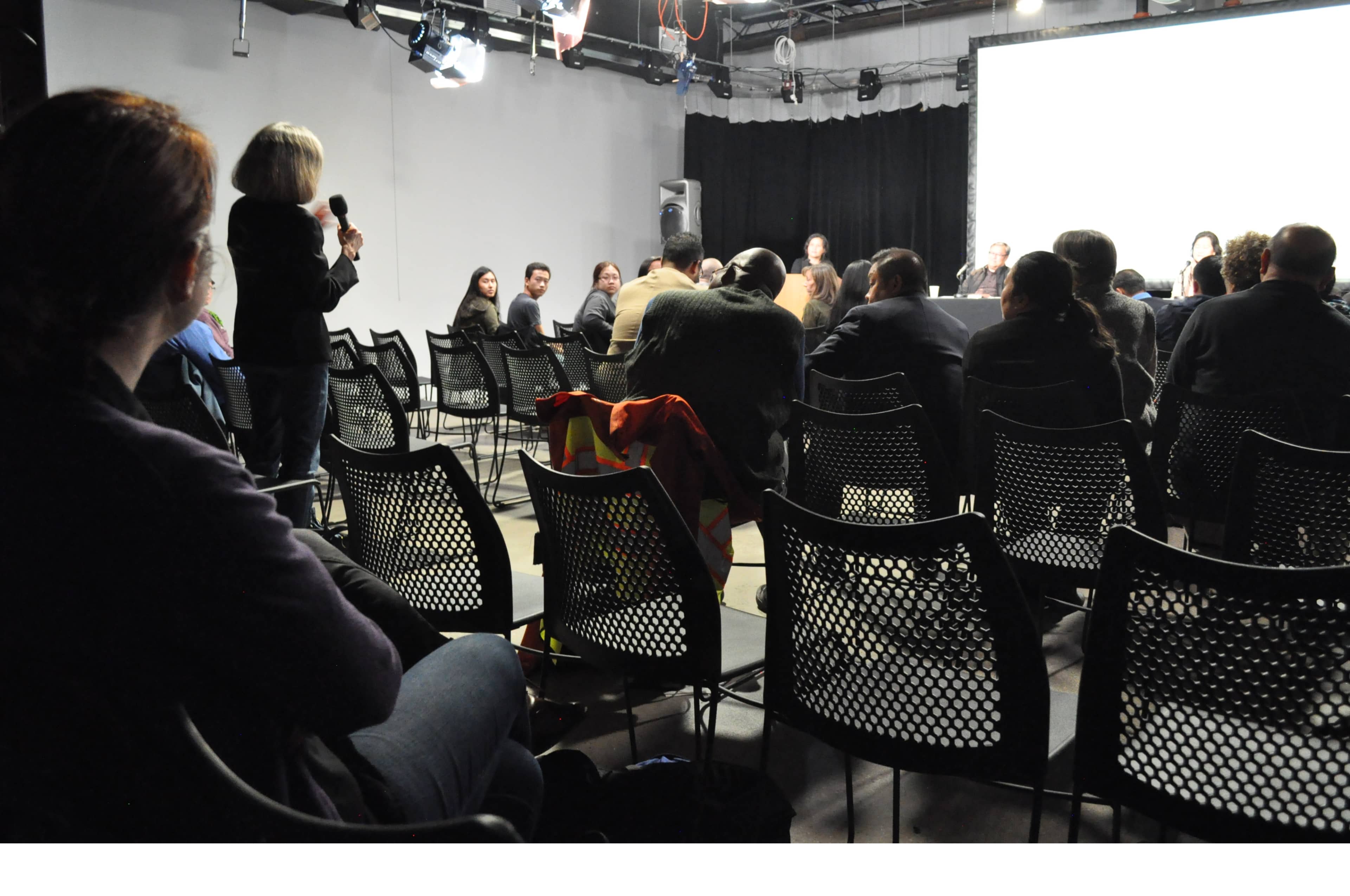 A woman holds a microphone while standing in front of a seated audience during a panel discussion in a dimly lit room.