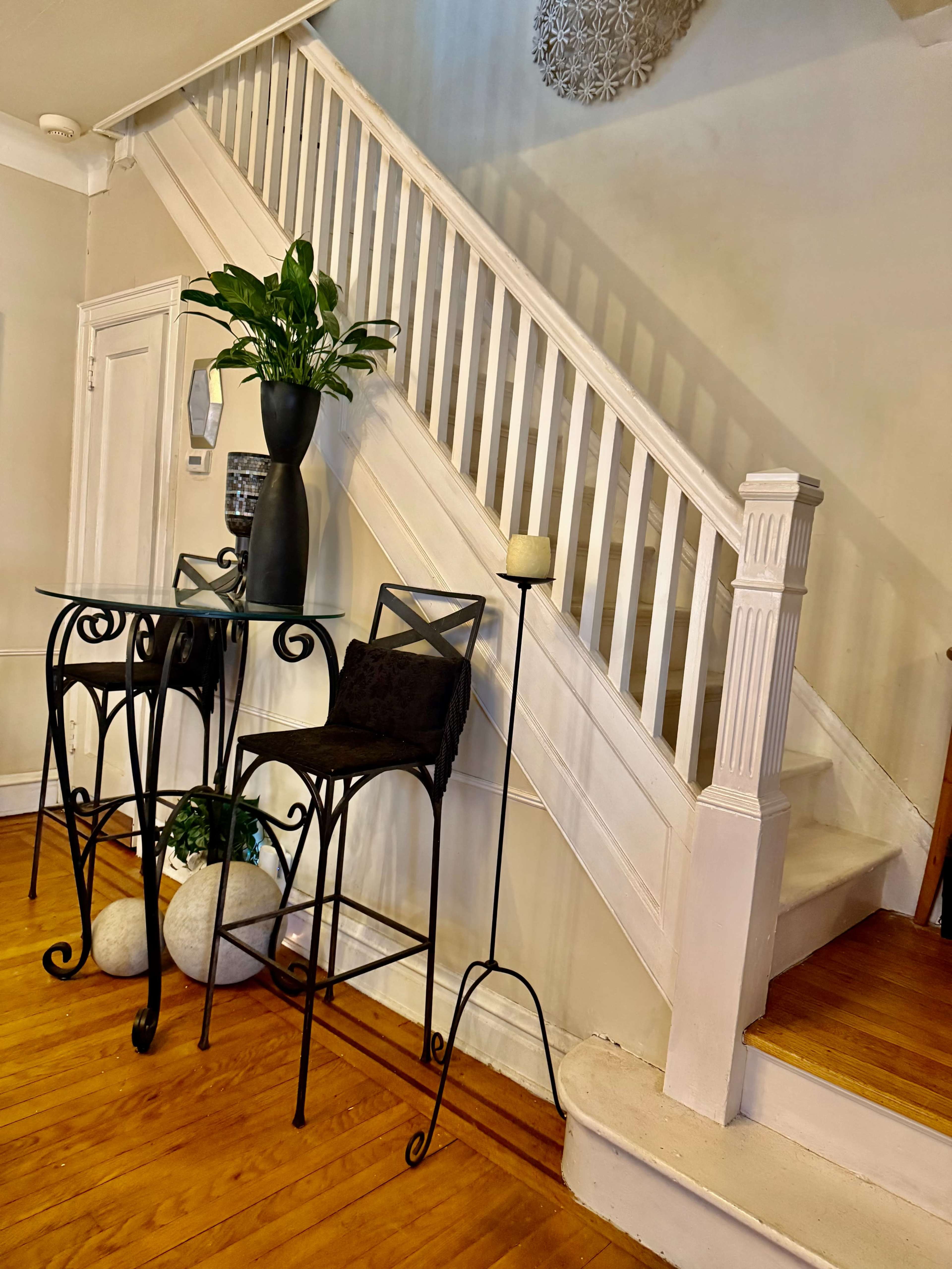 The image shows a staircase in a room with a small round table and two high-backed stools positioned beside it, along with a potted plant and a candle holder nearby.