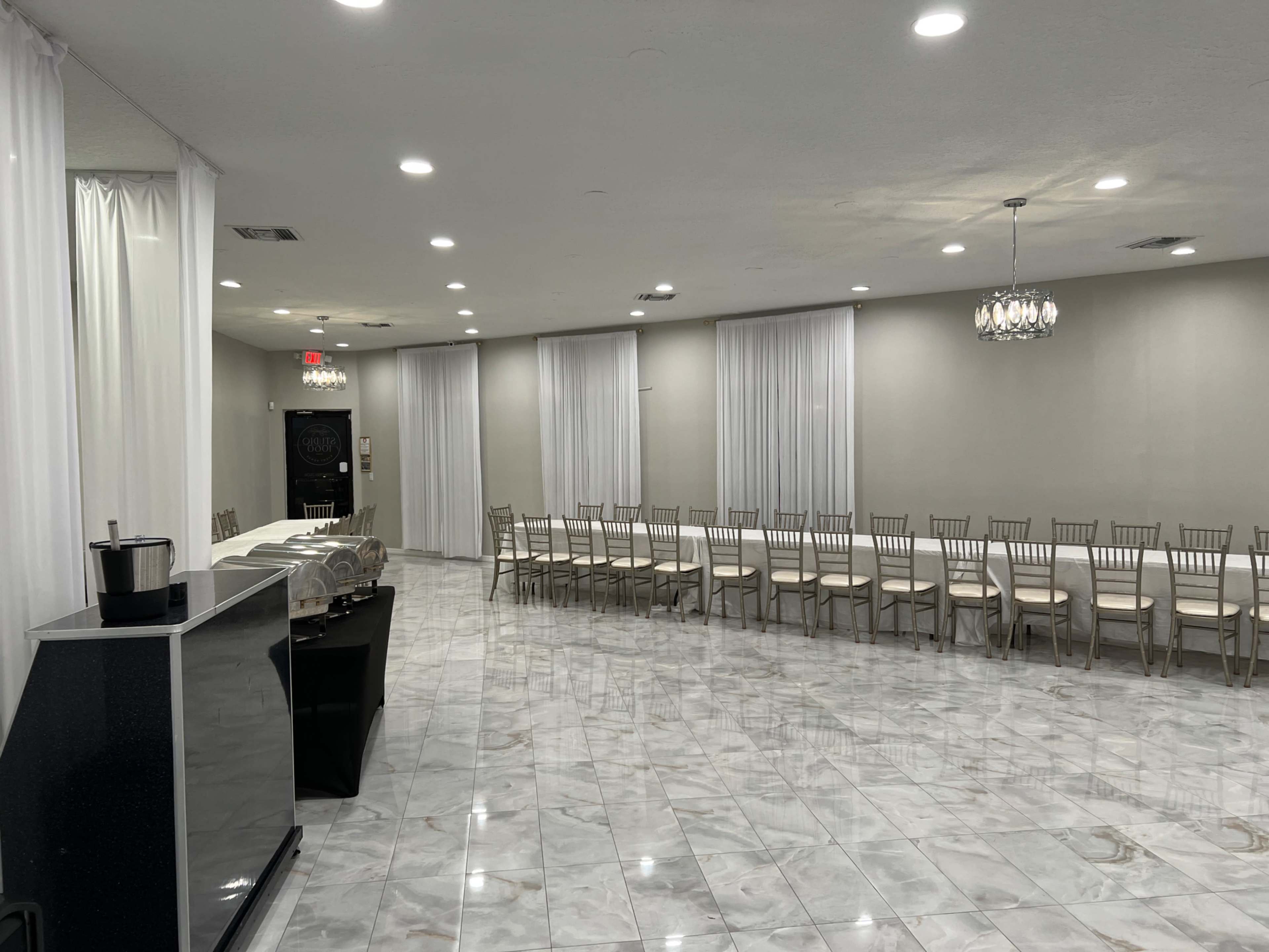 The image shows a spacious, well-lit room with white curtains, a long table with chairs arranged along its sides, and a bar area with a chilled beverage container.