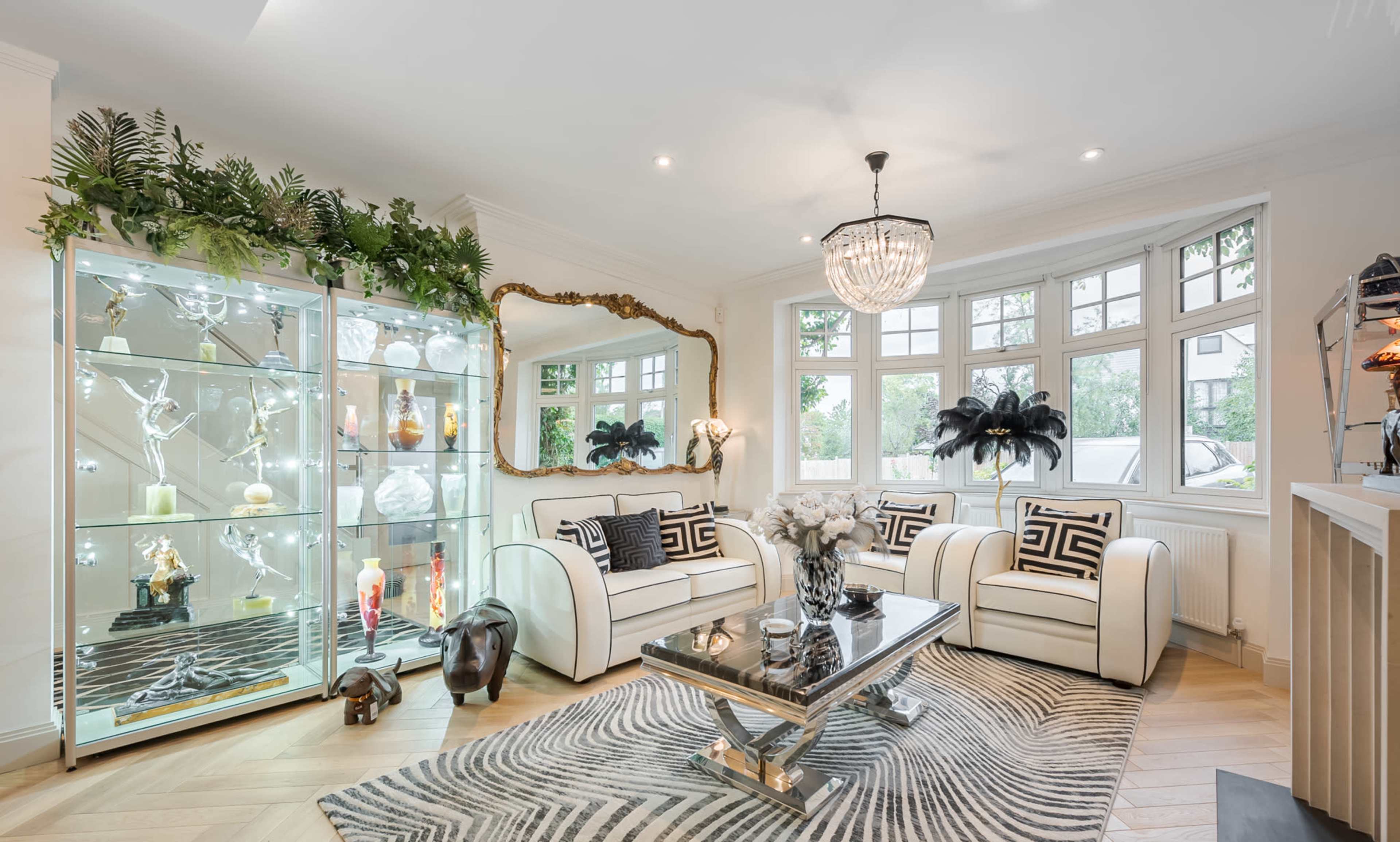A modern living room features two white sofas facing a glass display cabinet and a mirrored wall, with decorative plants and a chandelier overhead.