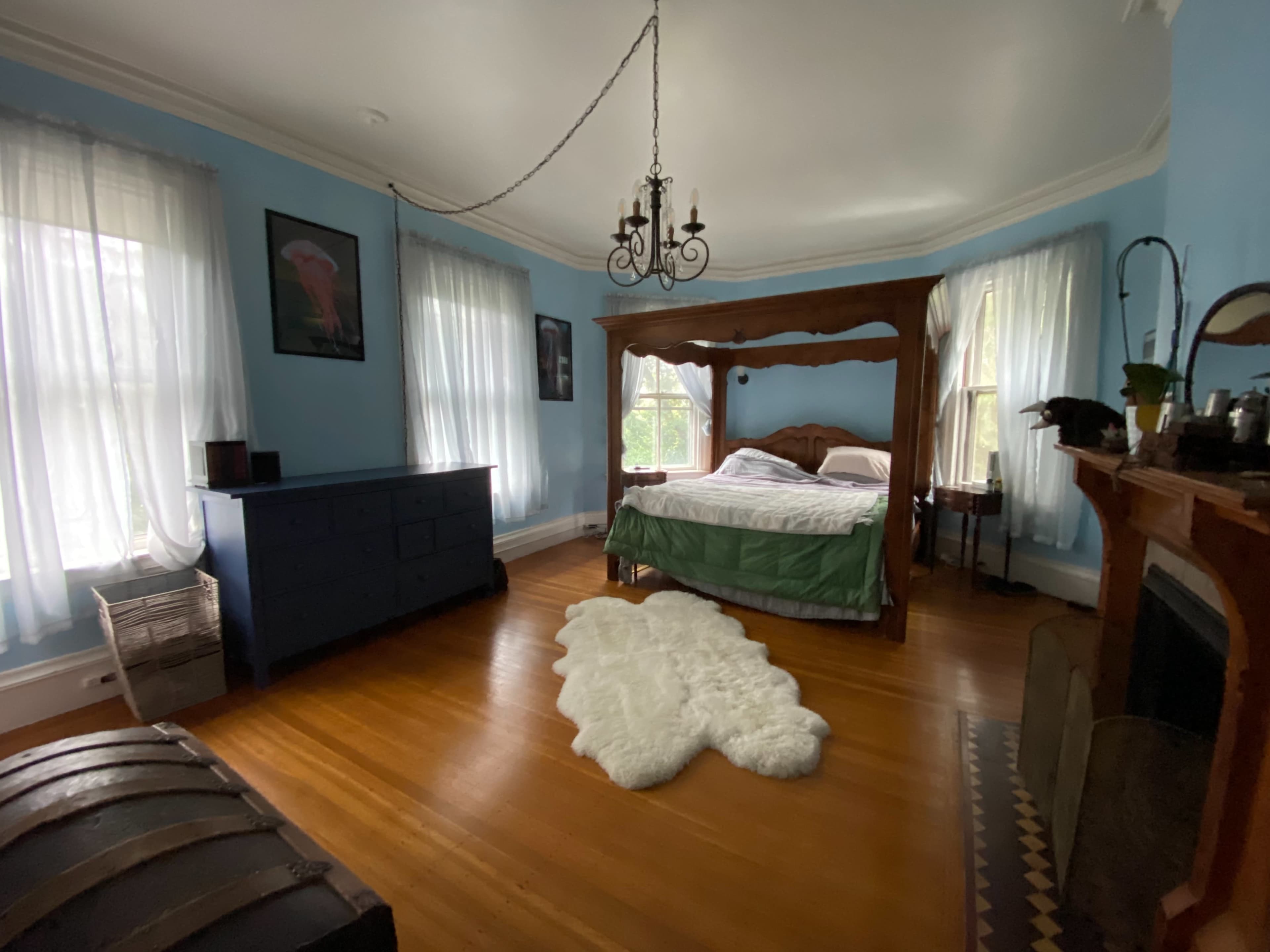 The room features a four-poster bed with a green bedspread, a blue dresser, and a white rug on a wooden floor, illuminated by natural light through sheer curtains.