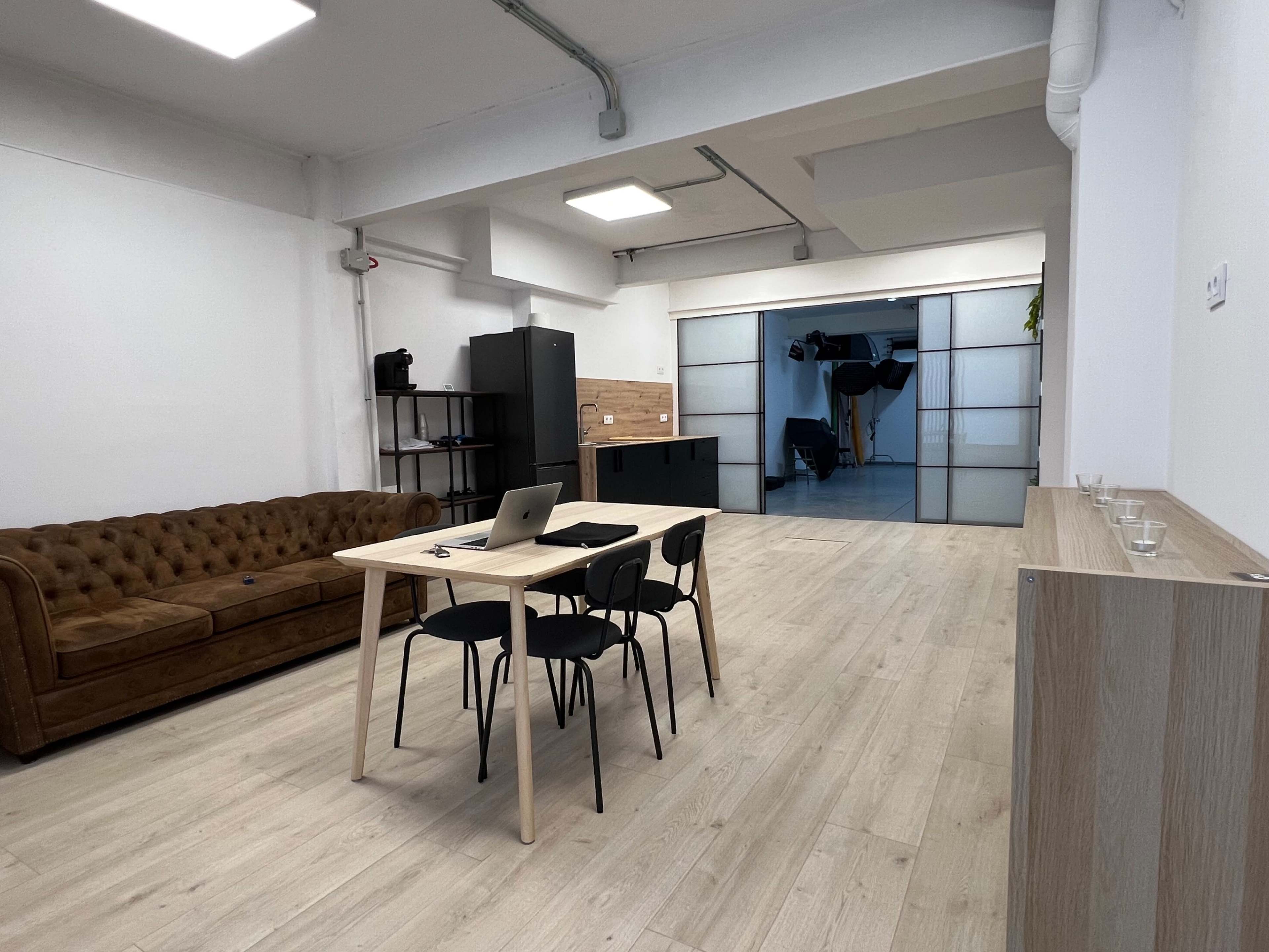 The image shows a spacious, modern office interior with a wooden table and chairs, a brown leather sofa, and glass partitioning leading to another area.