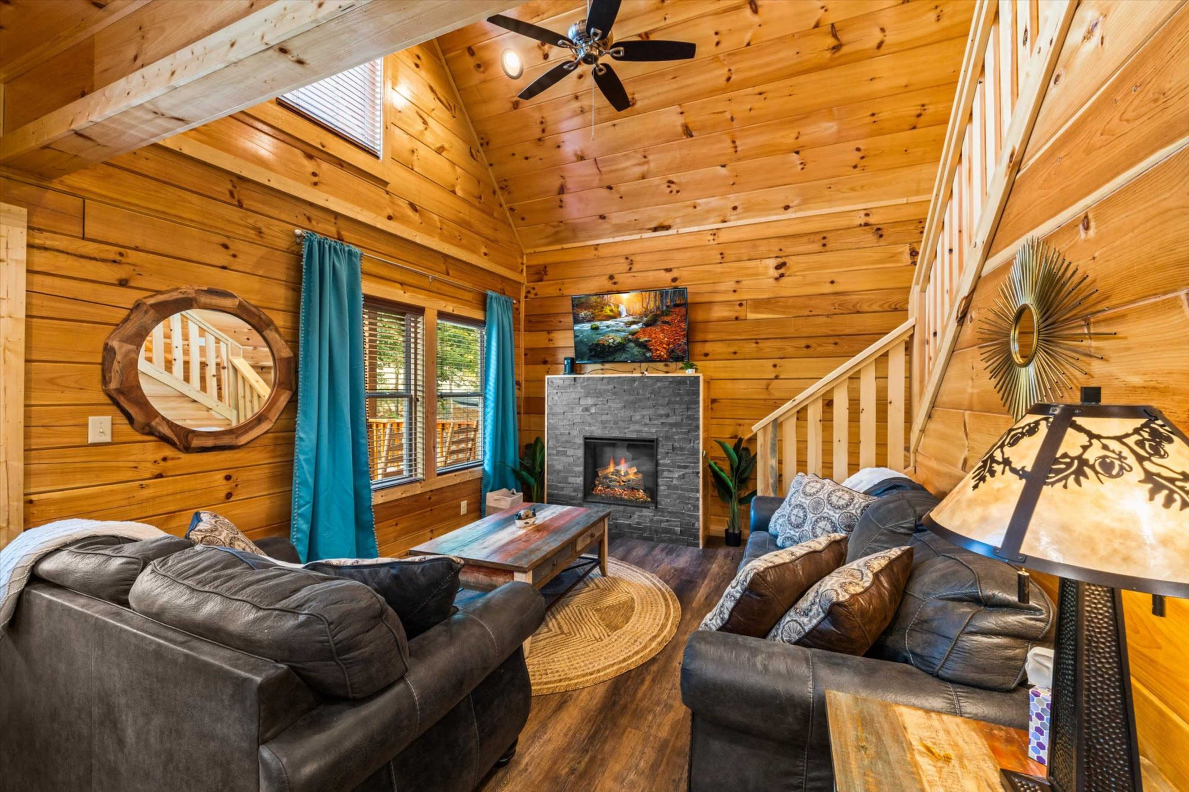 A cozy wooden cabin living room features a stone fireplace, two sofas, and large windows with blue curtains.