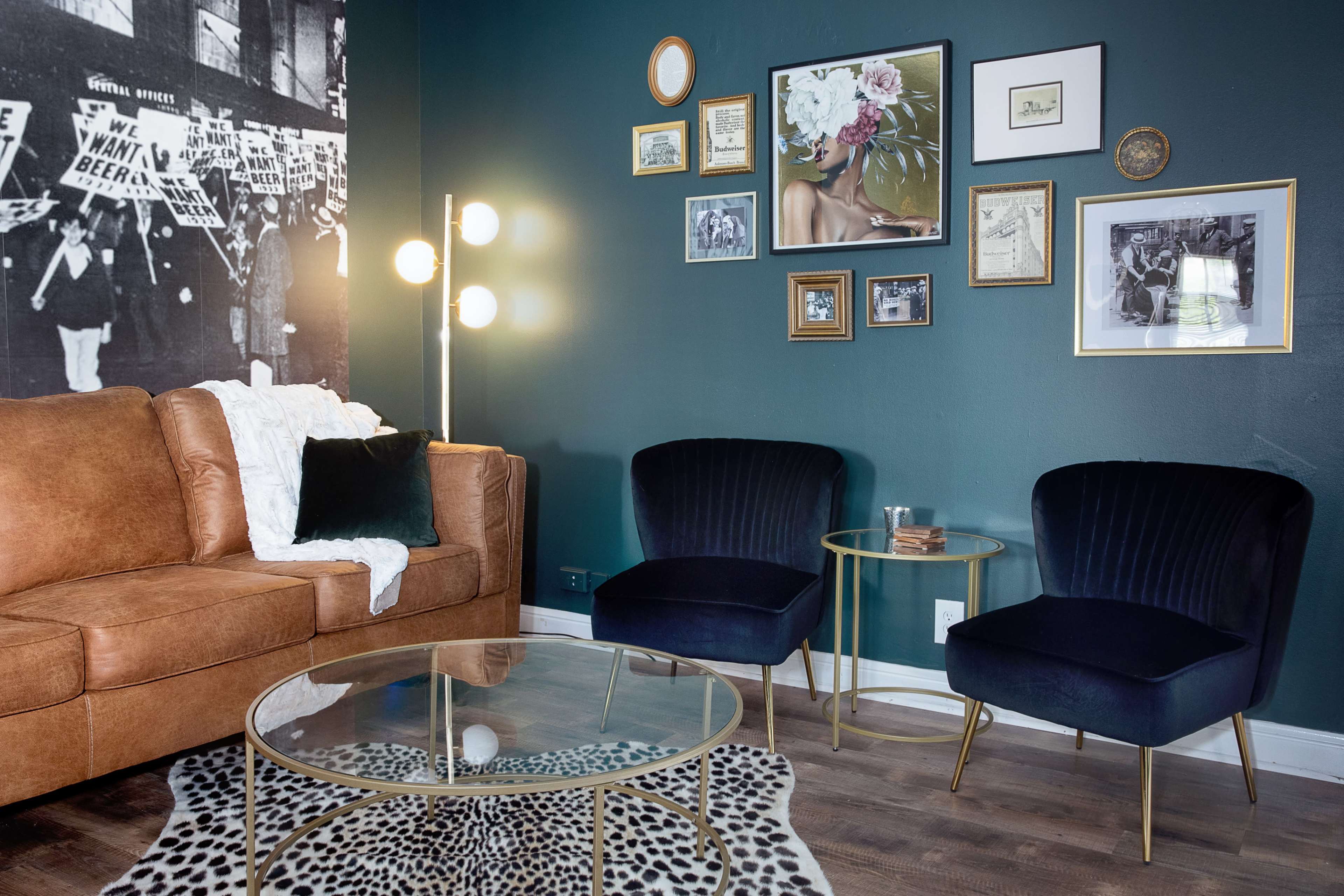 The image shows a stylish living room with a brown sofa, two navy armchairs, a glass coffee table, and a gallery wall of framed pictures against a dark green backdrop.