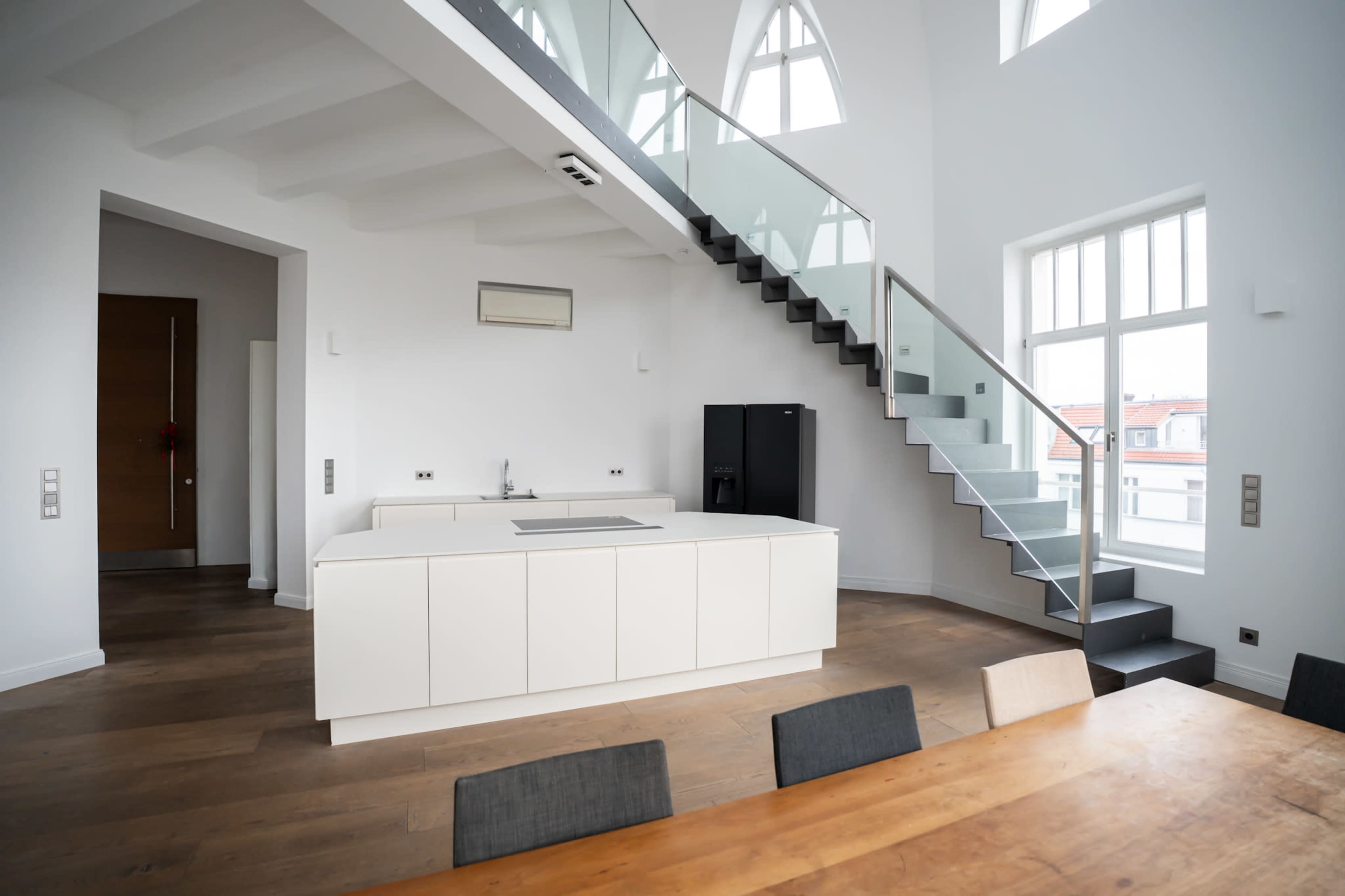 The image shows a modern, open-concept kitchen and dining area with a staircase and large windows letting in natural light.