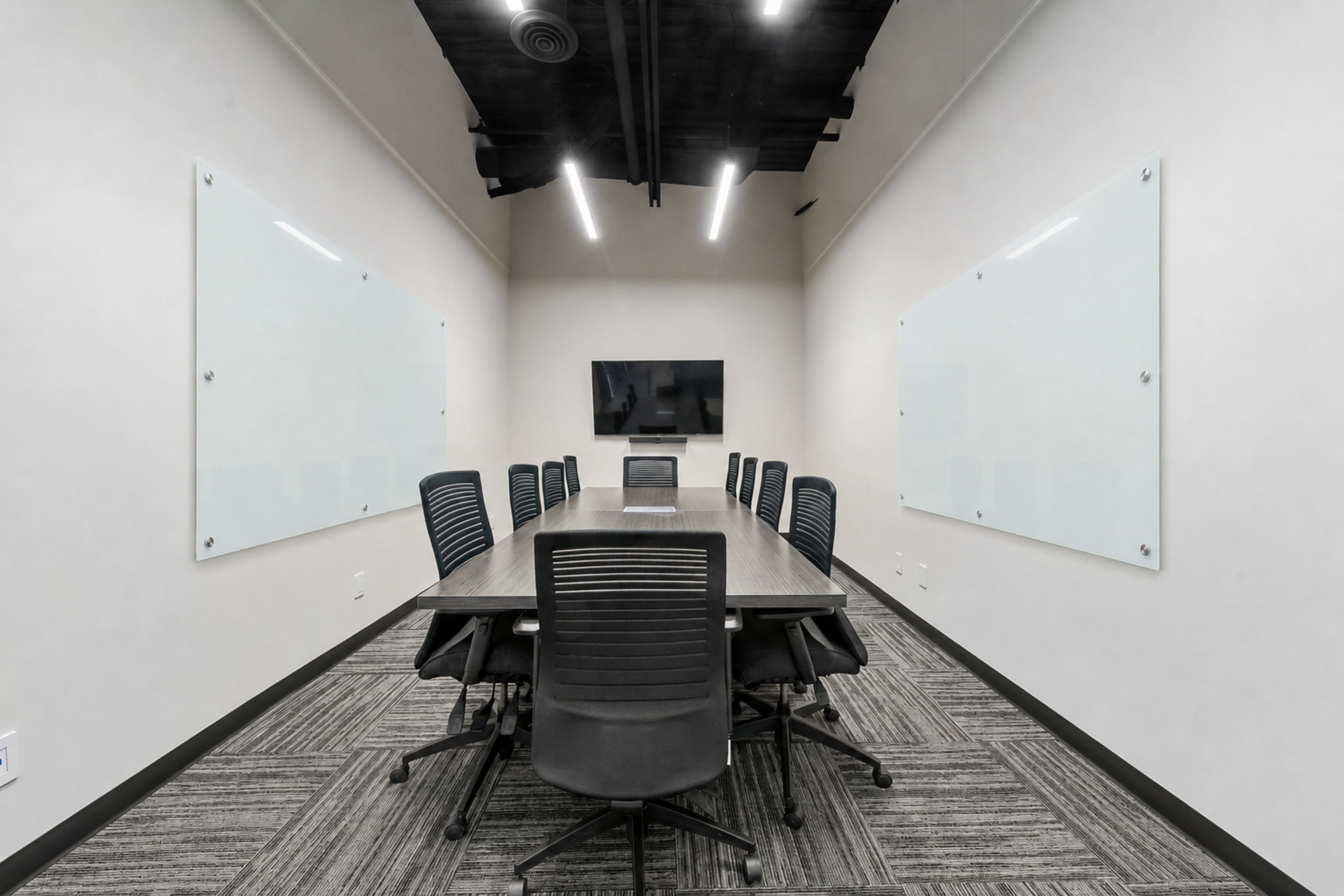 The image shows a modern conference room with a long table, multiple chairs, and a wall-mounted television surrounded by whiteboards.
