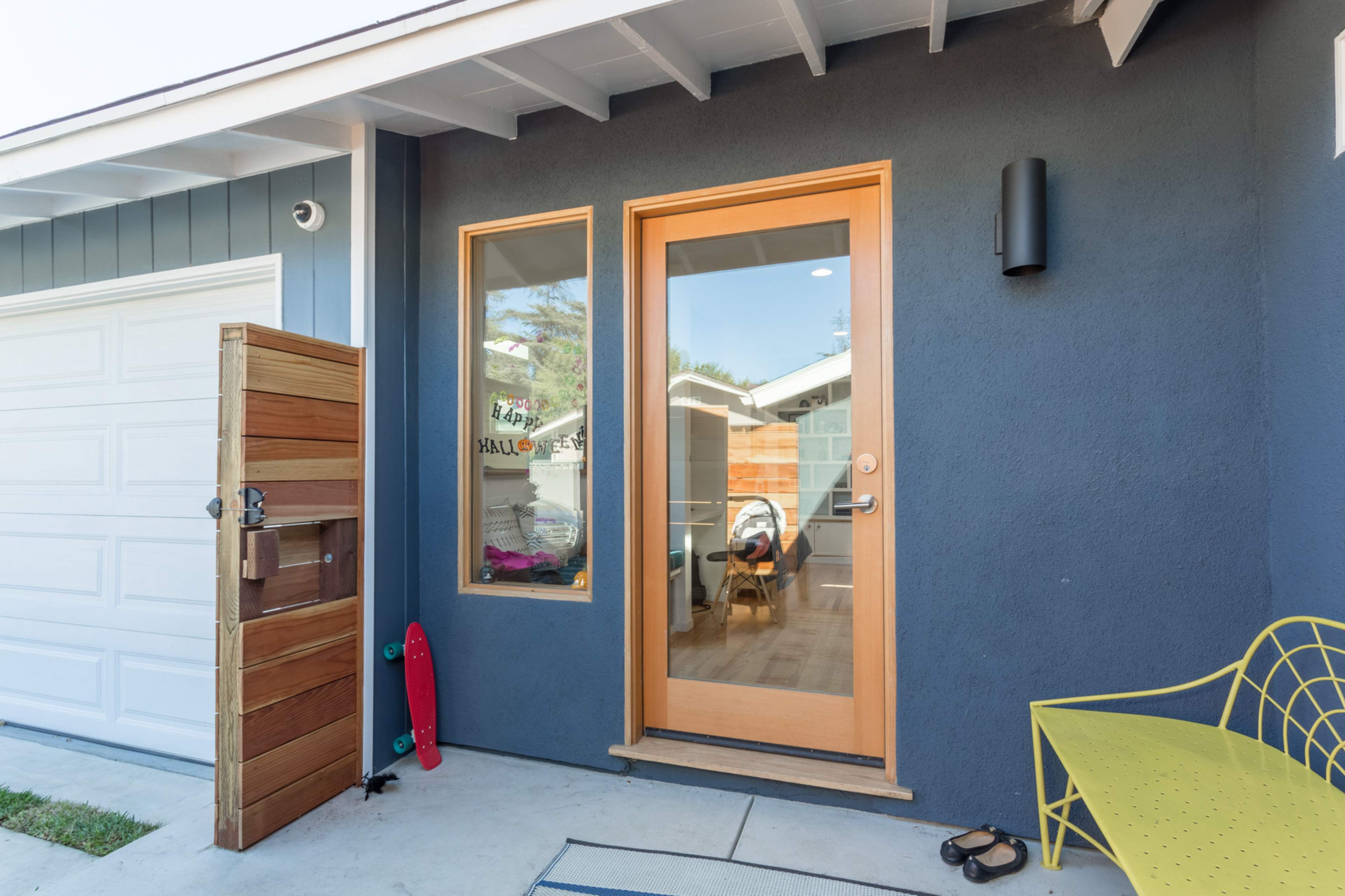 The image shows a modern front entrance with a wooden door, a decorative yellow chair, a skateboard leaning against the wall, and a wood-paneled mailbox beside it.