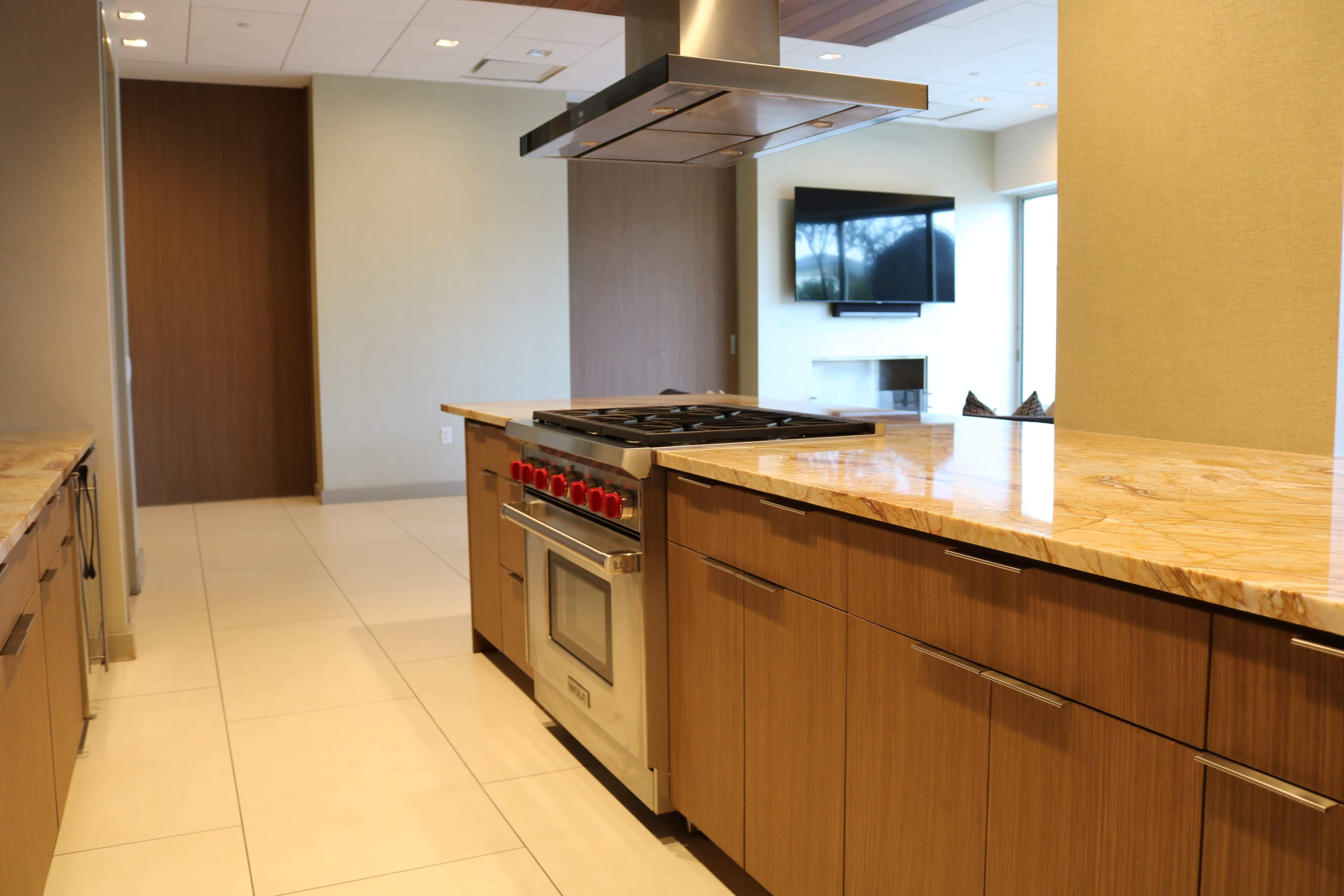 A modern kitchen featuring wooden cabinetry, a gas stove with red knobs, and a spacious countertop alongside a living area with a television and a fireplace.