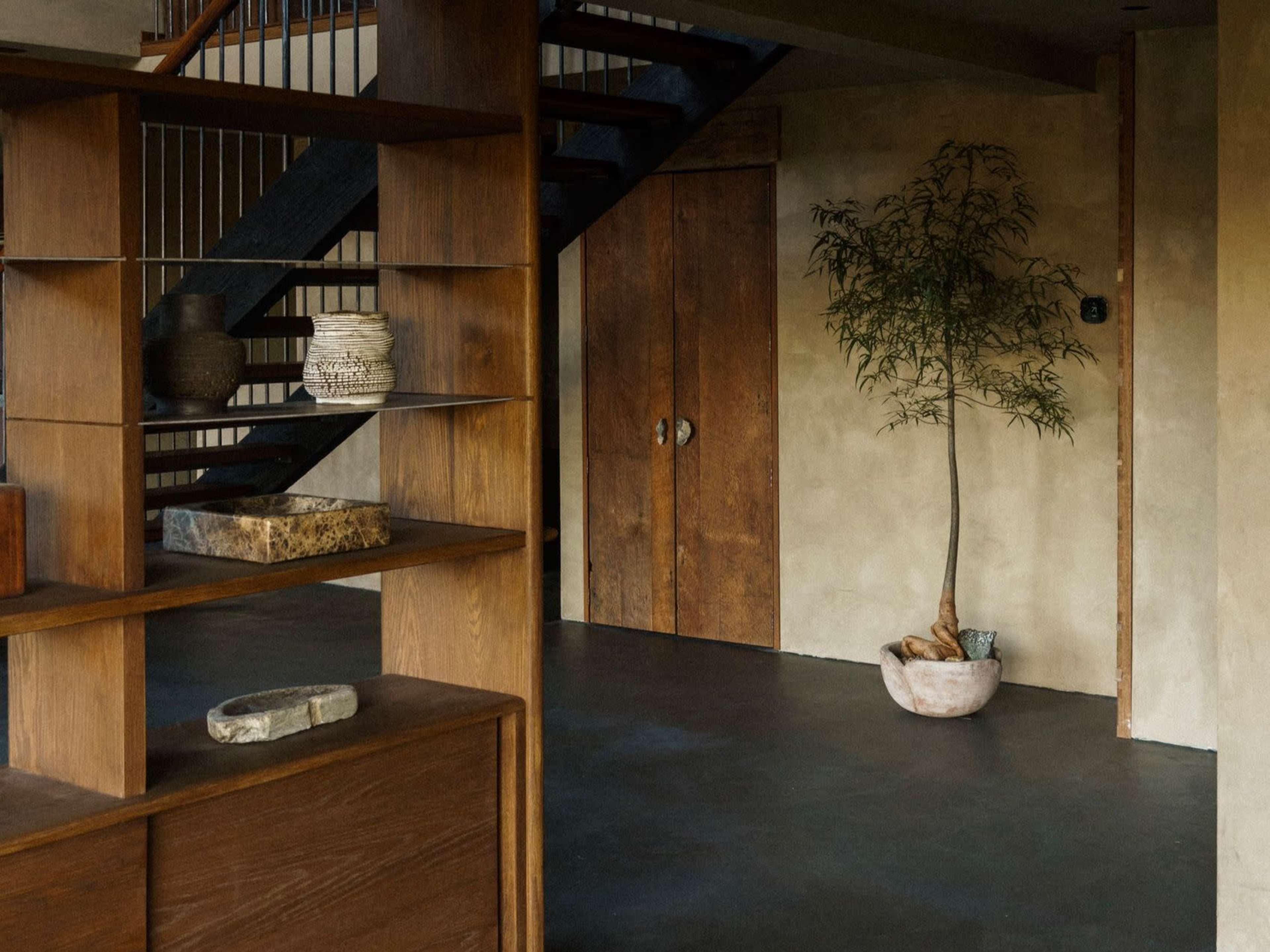 The image shows an interior scene featuring a staircase, wooden shelves with decorative items, and a potted plant by a textured wall.