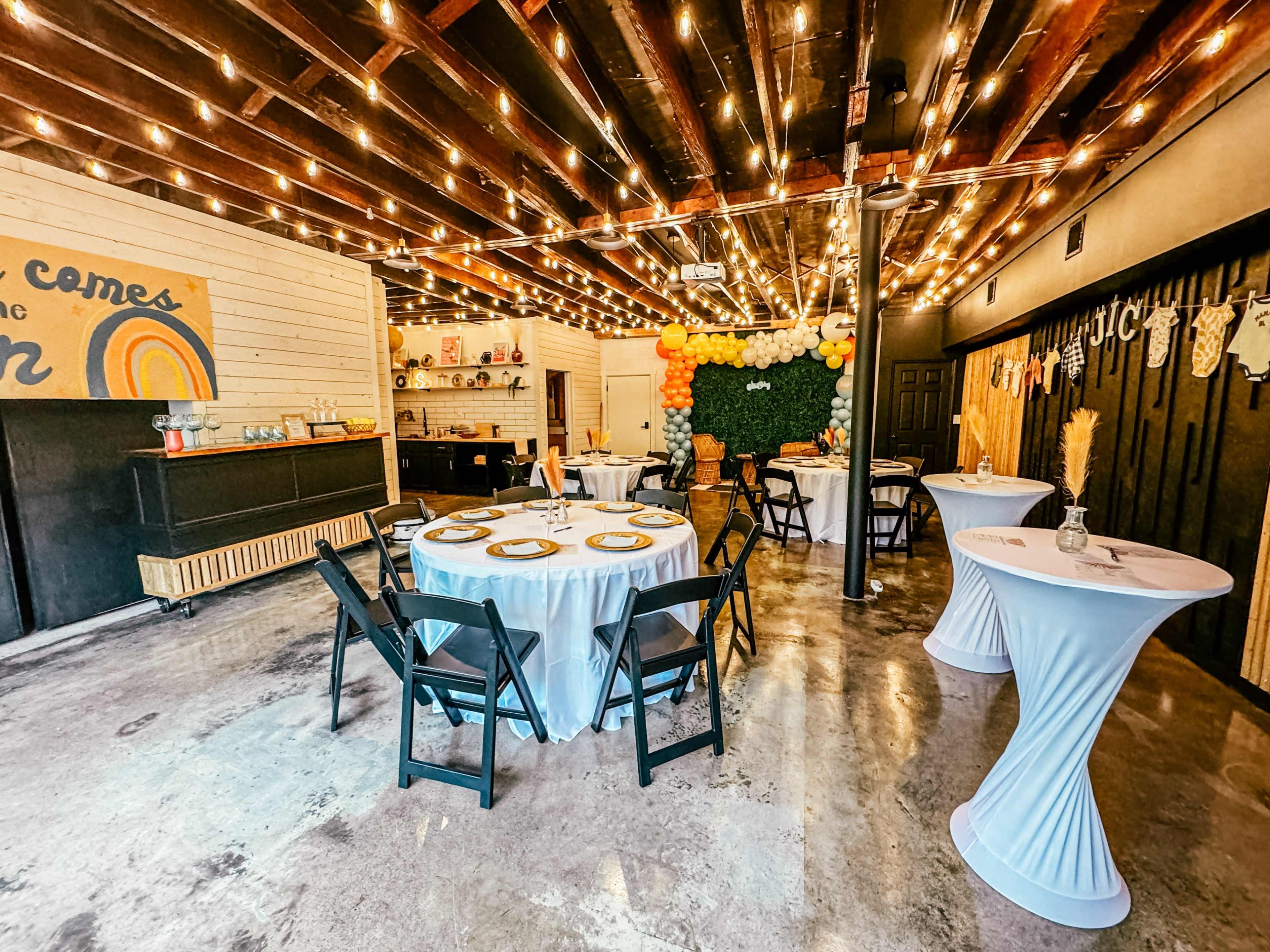 An event space decorated with string lights, featuring round tables with white tablecloths and a colorful balloon backdrop.