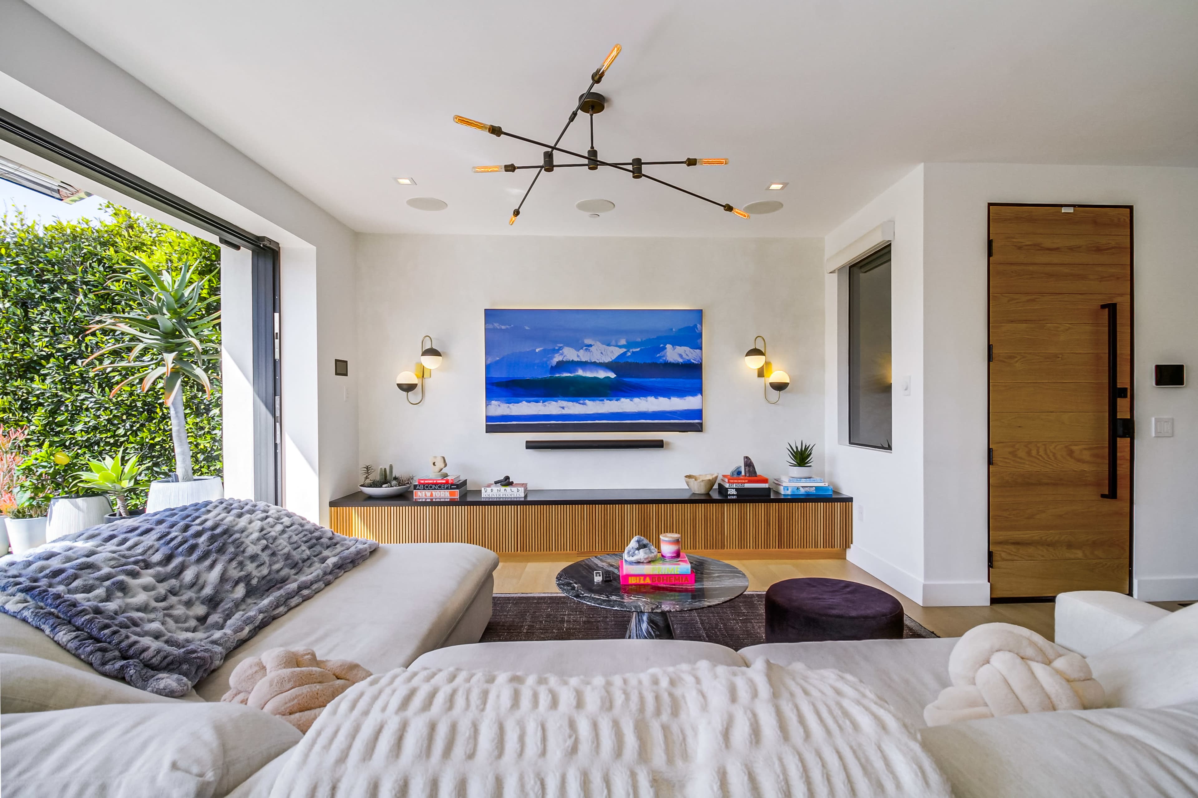 A modern living room features a large painting of ocean waves above a sleek wooden console, with a cozy seating arrangement and natural light streaming in through large sliding doors.