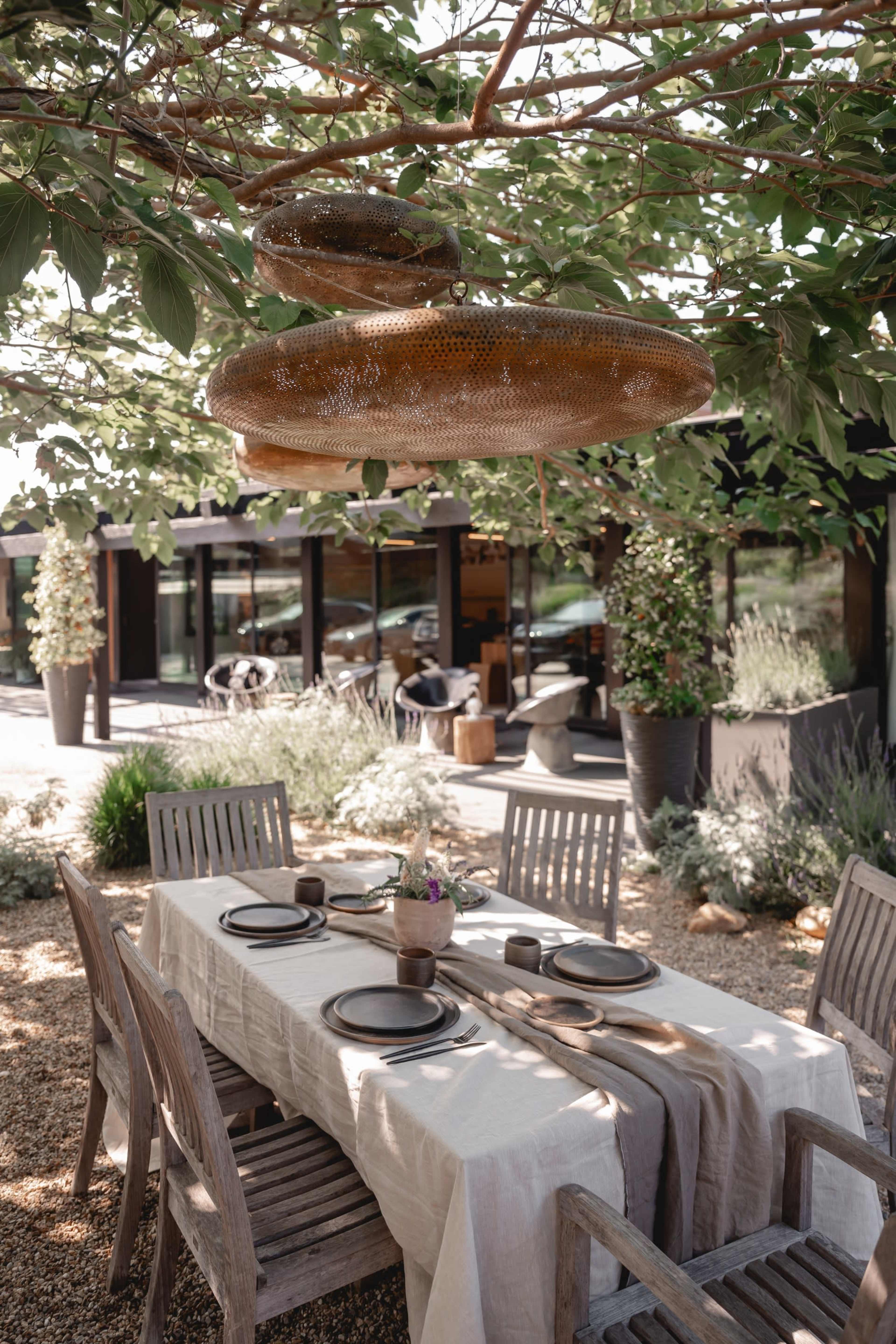 A wooden dining table is set under a tree with hanging light fixtures, surrounded by plants and a modern building in the background.