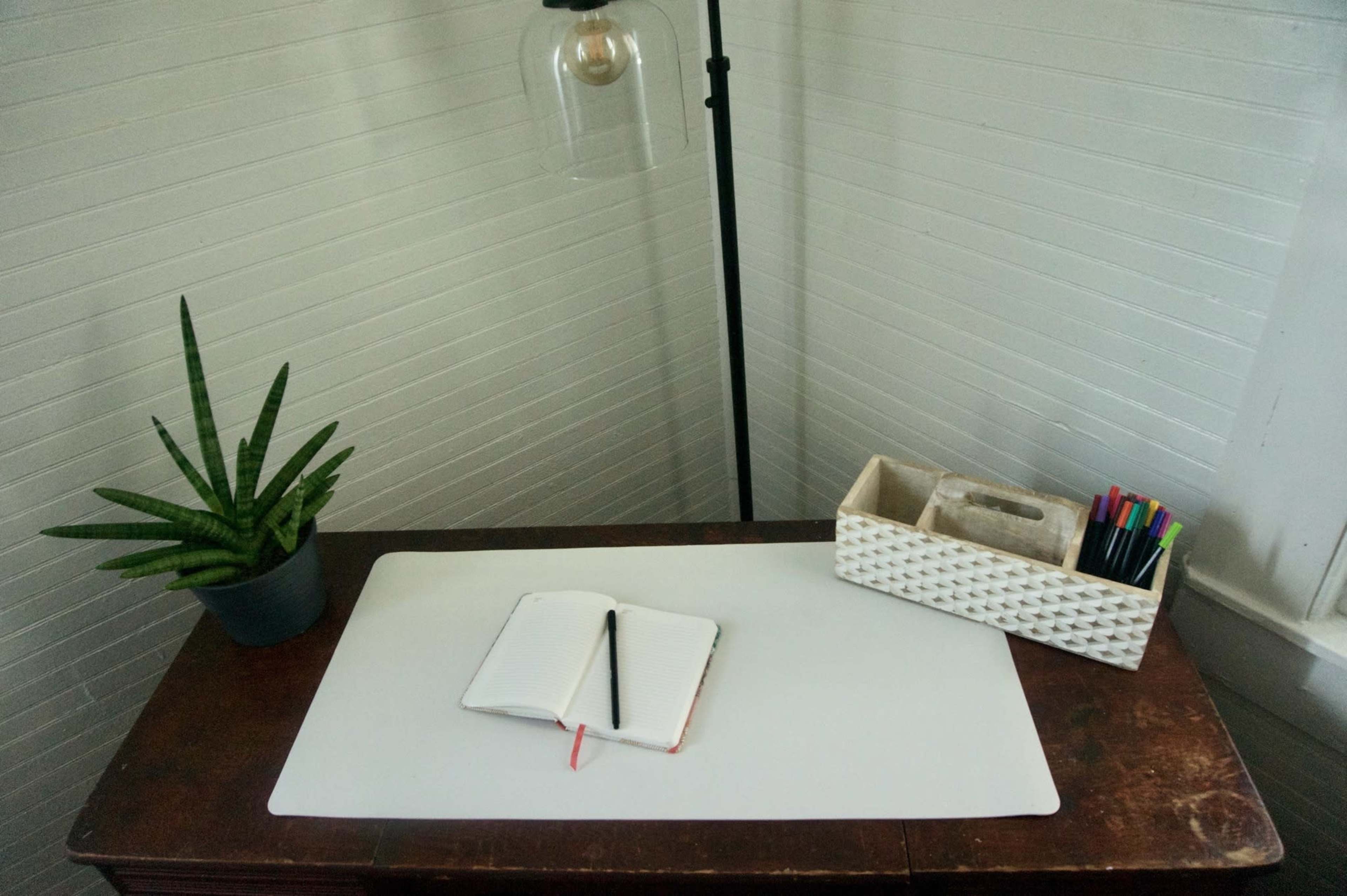 A wooden desk is displayed with a notebook and pen, a potted plant, a lamp, and a container of markers.