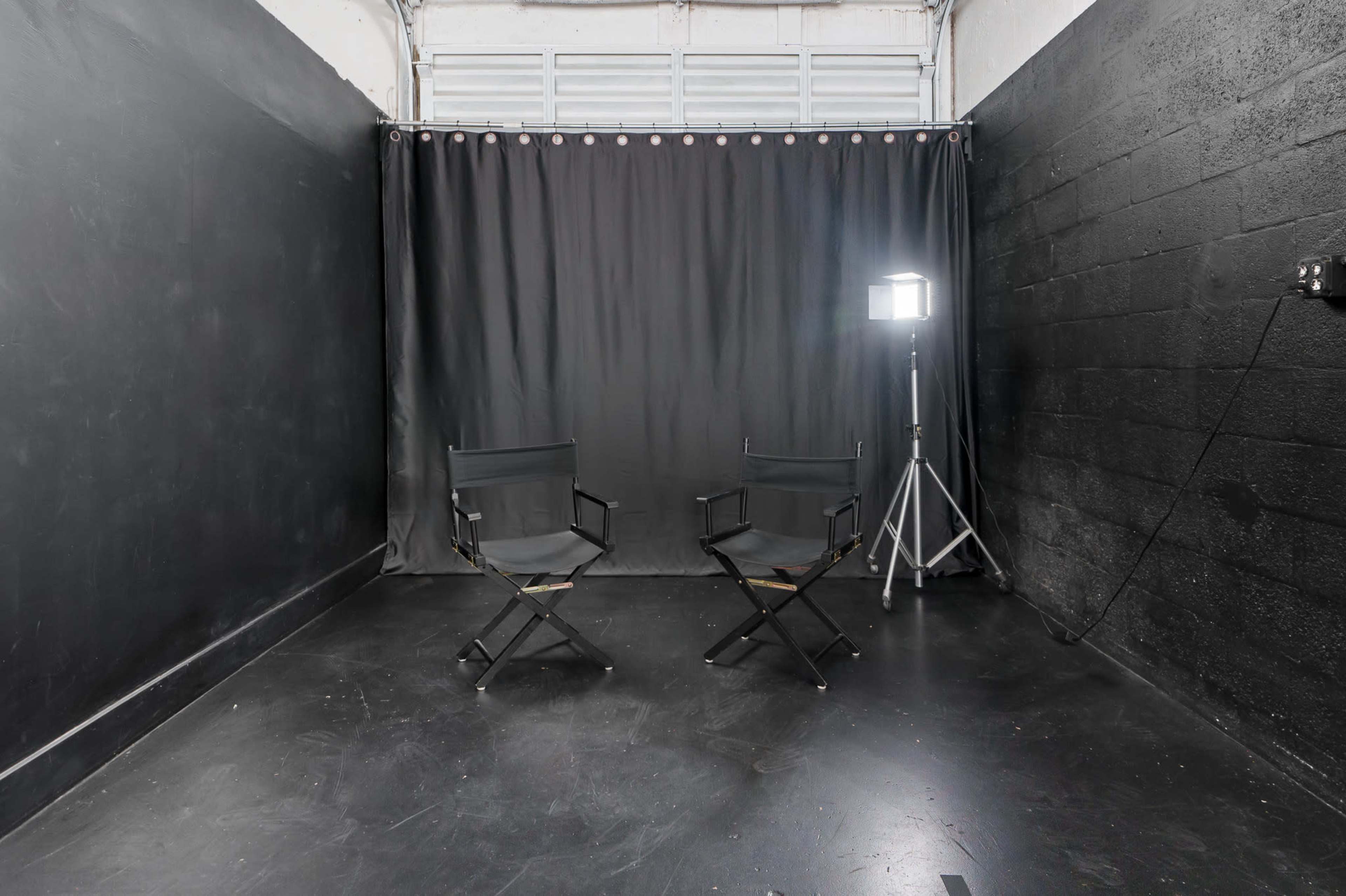 A dimly lit room with black walls and a black curtain, featuring two director's chairs and a floor lamp on a tripod.
